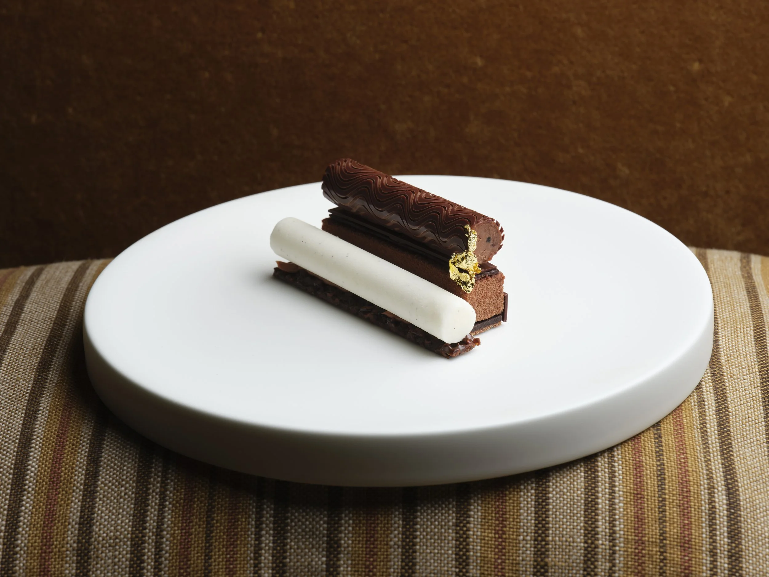 A modern chocolate dessert with layered textures, topped with wavy chocolate glaze, gold leaf, and served on a white plate with a brown background.