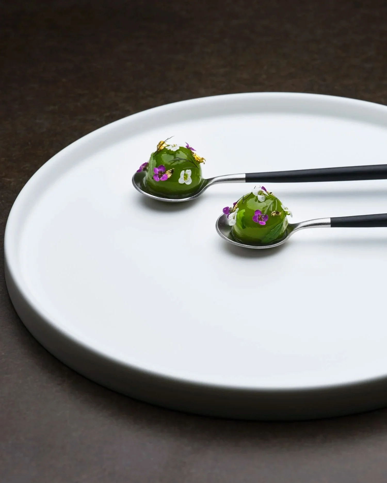 Two silver spoons with green jelly topped with edible flowers on a white plate.