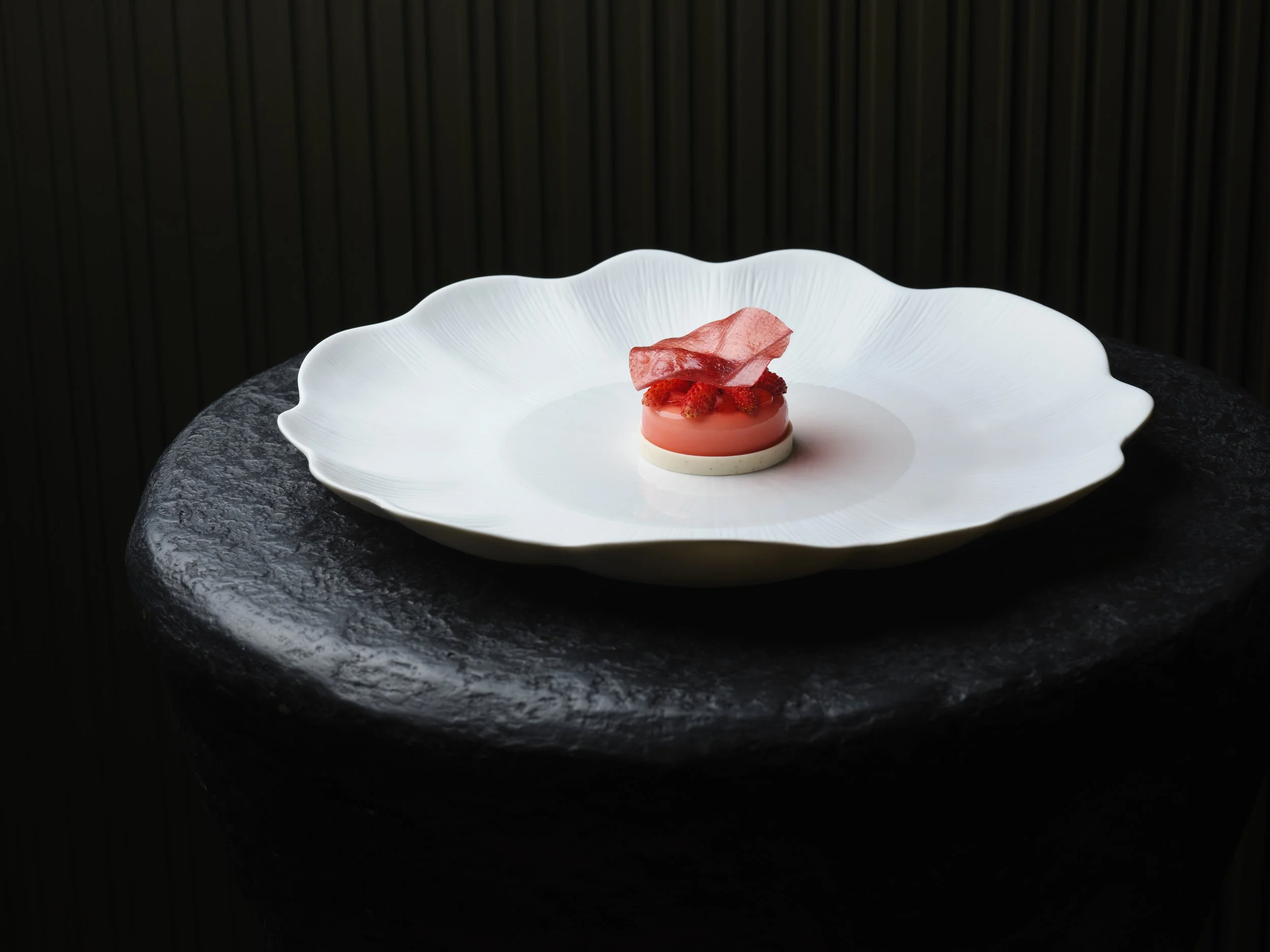A white decorative bowl with fluted edges containing a English strawberry dessert garnished with berries and a translucent pink edible tuile, set on a dark textured surface with a black background of Woven Restaurant.