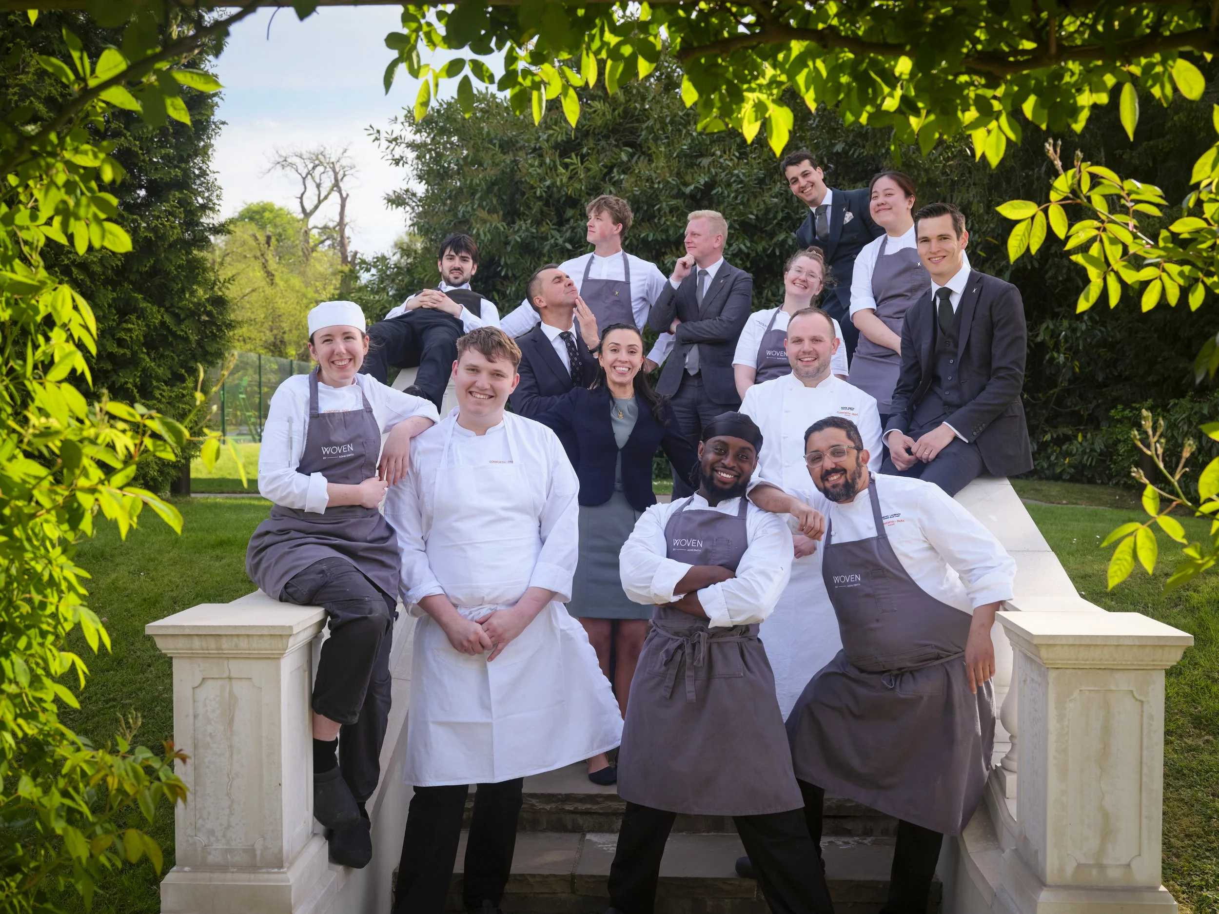 Group of diverse restaurant staff members outdoors, dressed in professional uniforms, smiling and posing together on a stone staircase with green trees in the background.