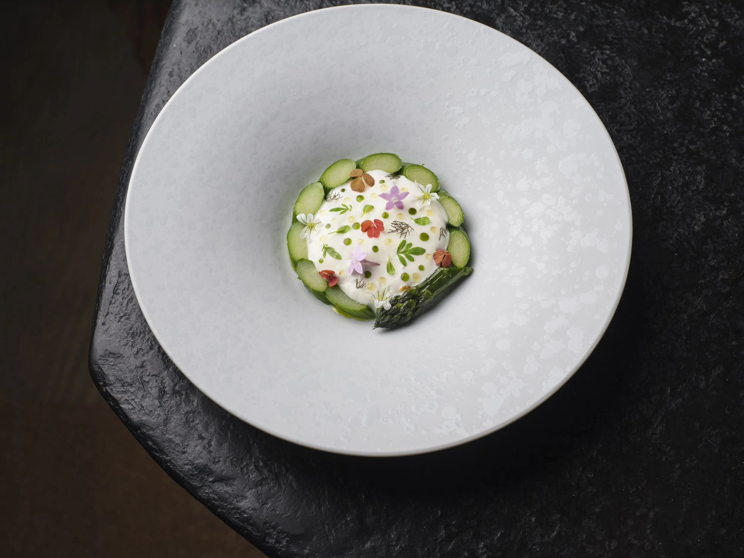 A plated dish of asparagus, sauce topped with edible flowers, asparagus slices, and herbs, garnished with small green and red leaves, set on a white plate on a dark textured surface.