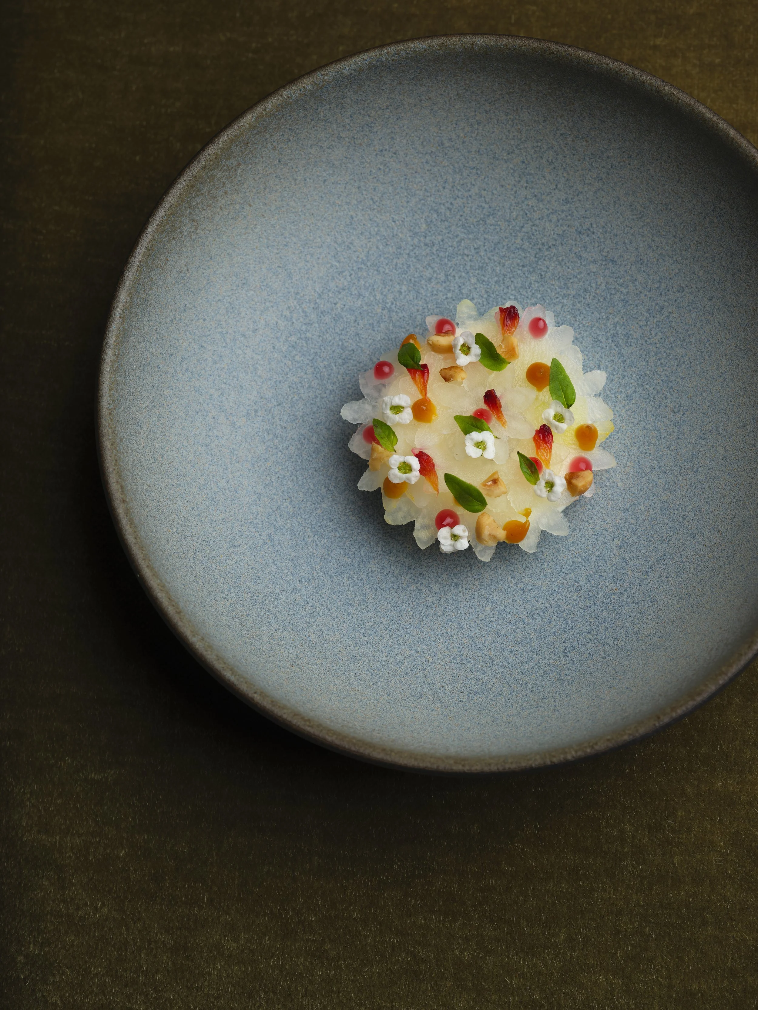 A blue ceramic plate with a small, colorful mound of finely chopped vegetables and herbs, garnished with edible flowers, herbs, and tiny decorative sauces on a dark surface.