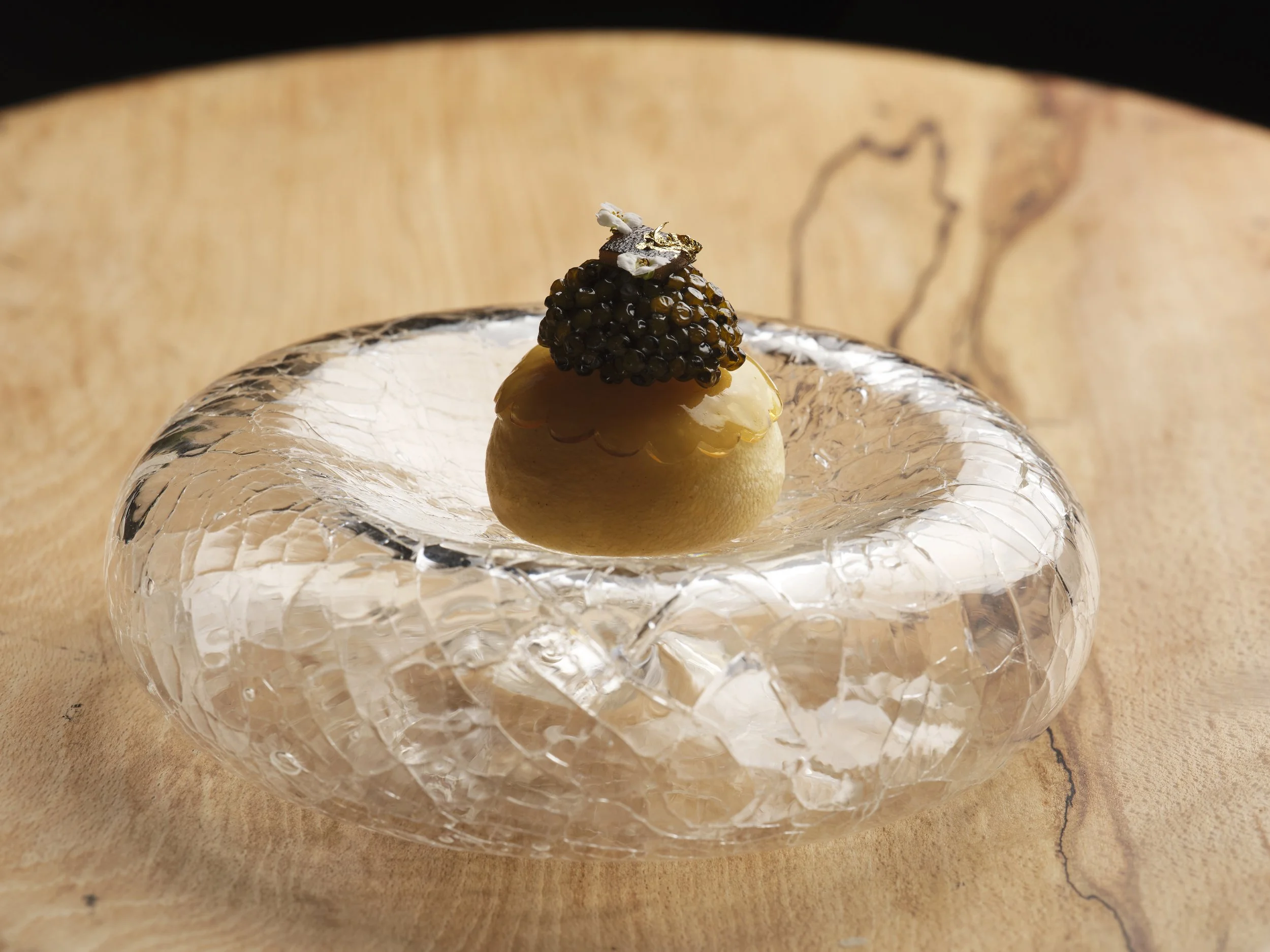 Close-up of a dome-shaped dessert topped with a cluster of black pearls, small white edible flowers, and a piece of gold leaf, placed on a clear textured glass plate on a wooden surface.