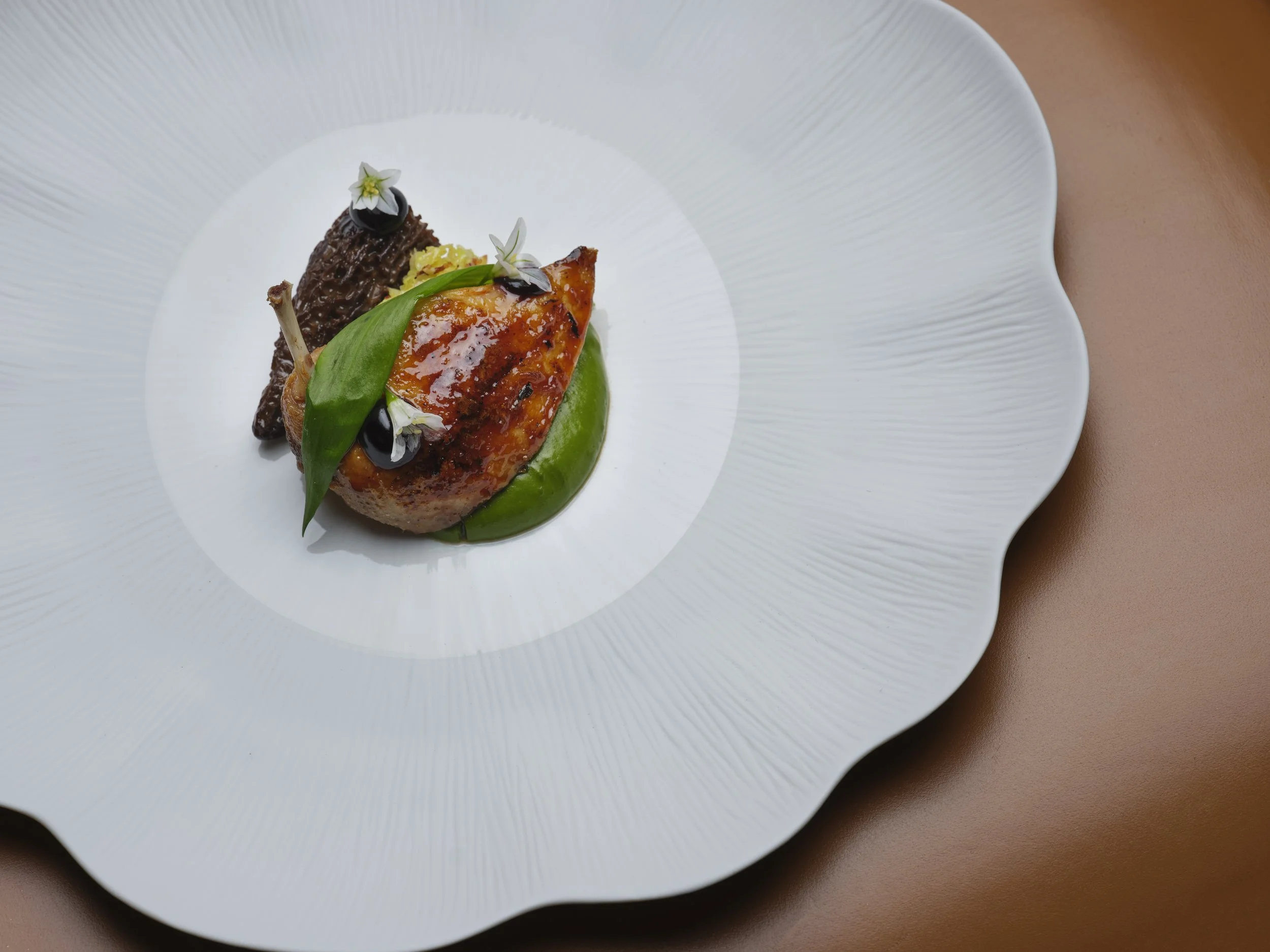 Close-up of a plated roasted qual with a glazed piece of meat, green sauce, a leaf, small edible flowers, and chocolate garnishes on a white textured plate.