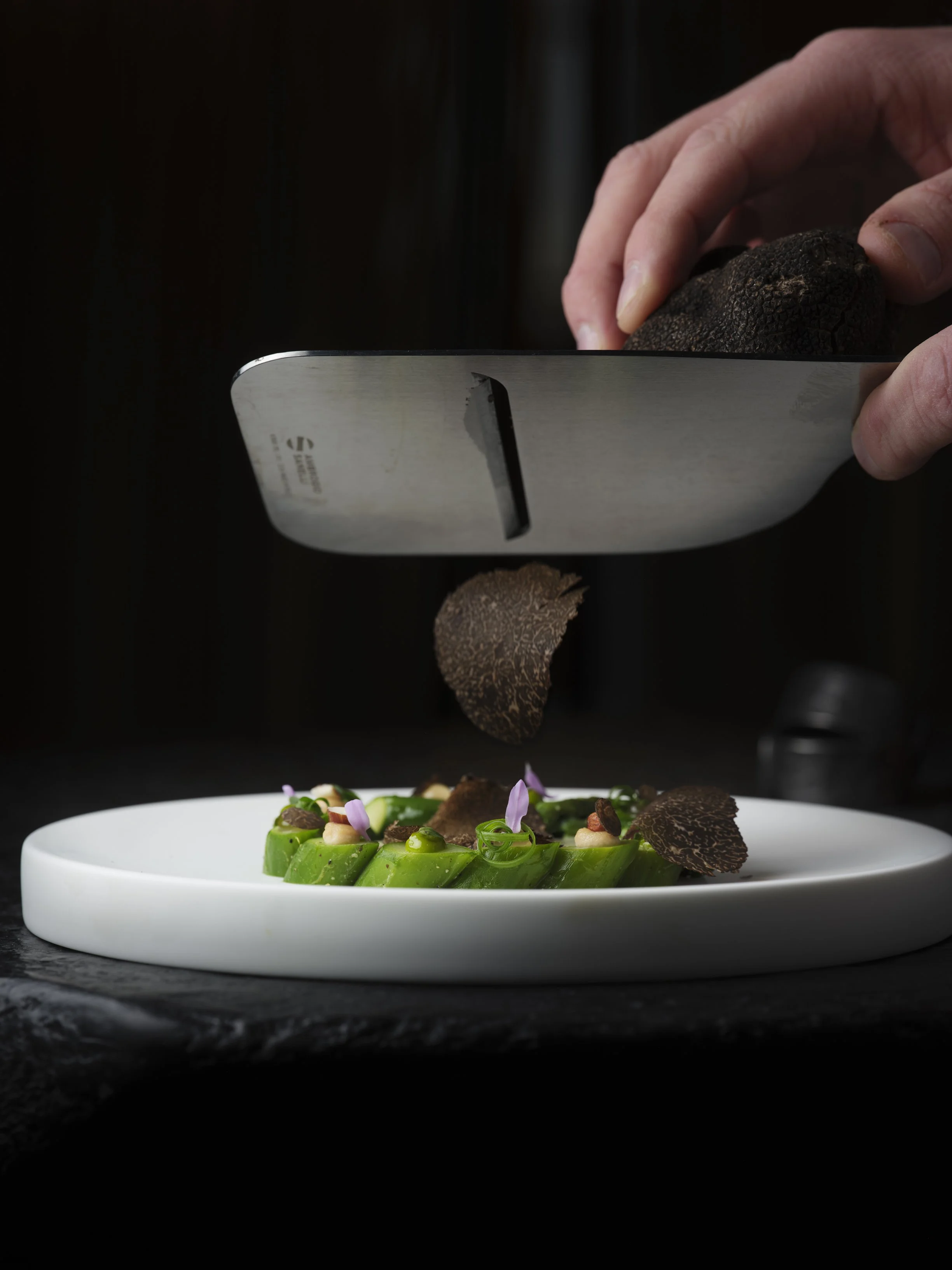 Chef using a grater to shave black truffle over a dish of sliced cucumber garnished with herbs and edible flowers on a white plate.