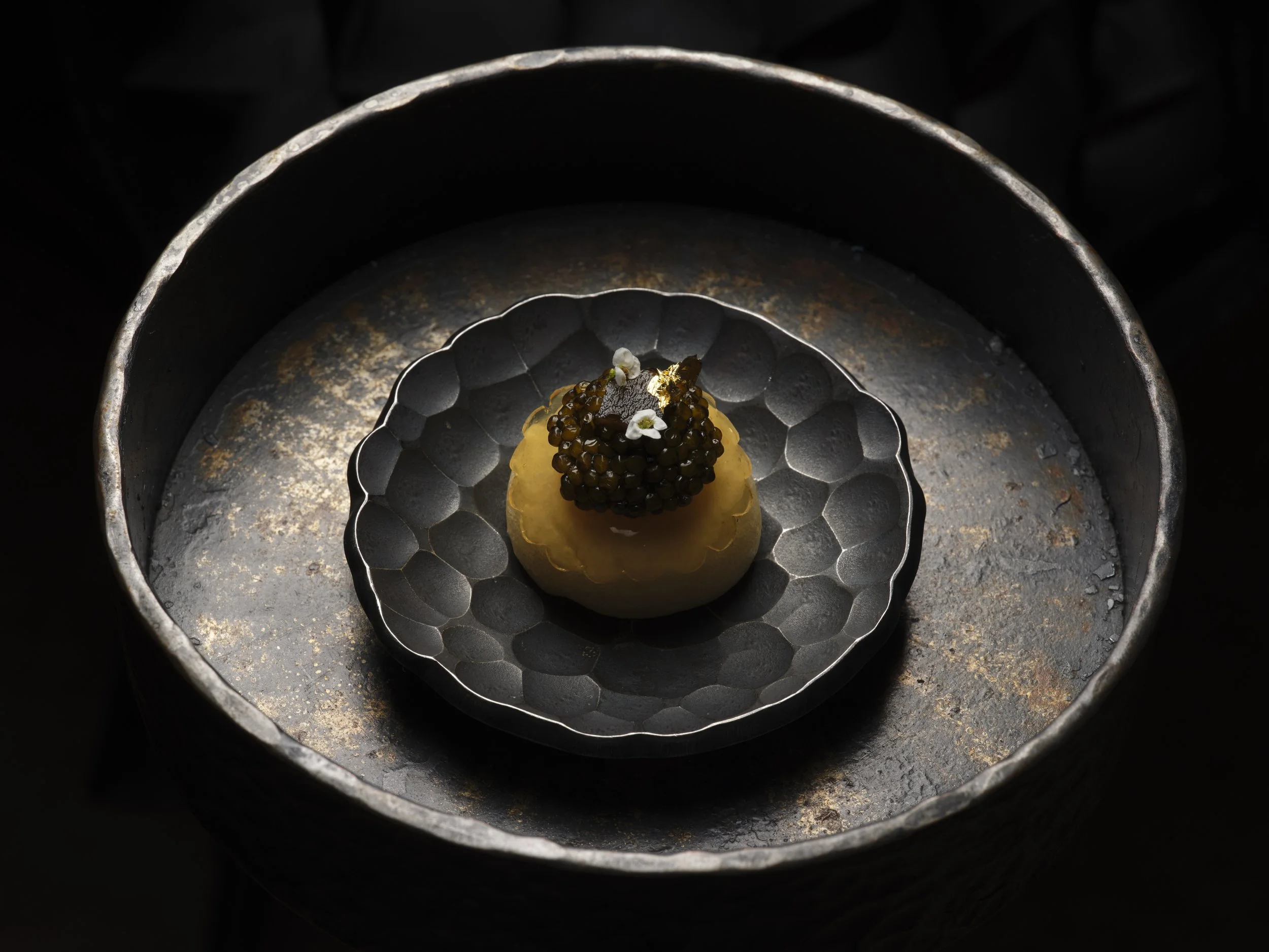 Close-up of a dessert with black caviar-like toppings, garnished with small white edible flowers, served on a dark textured surface with concentric circular patterns.