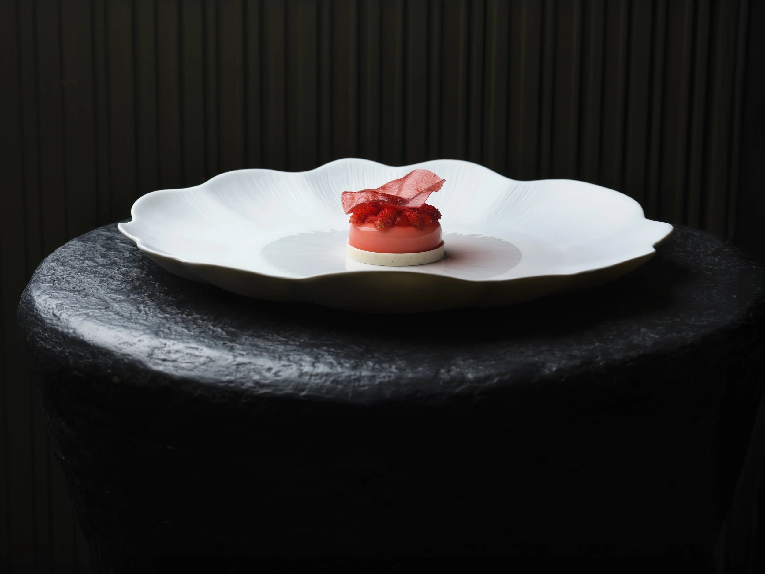 A decorative dessert with pink glaze, topped with red berries and a pink decorative piece, served on a white wavy-edge plate against a dark background.