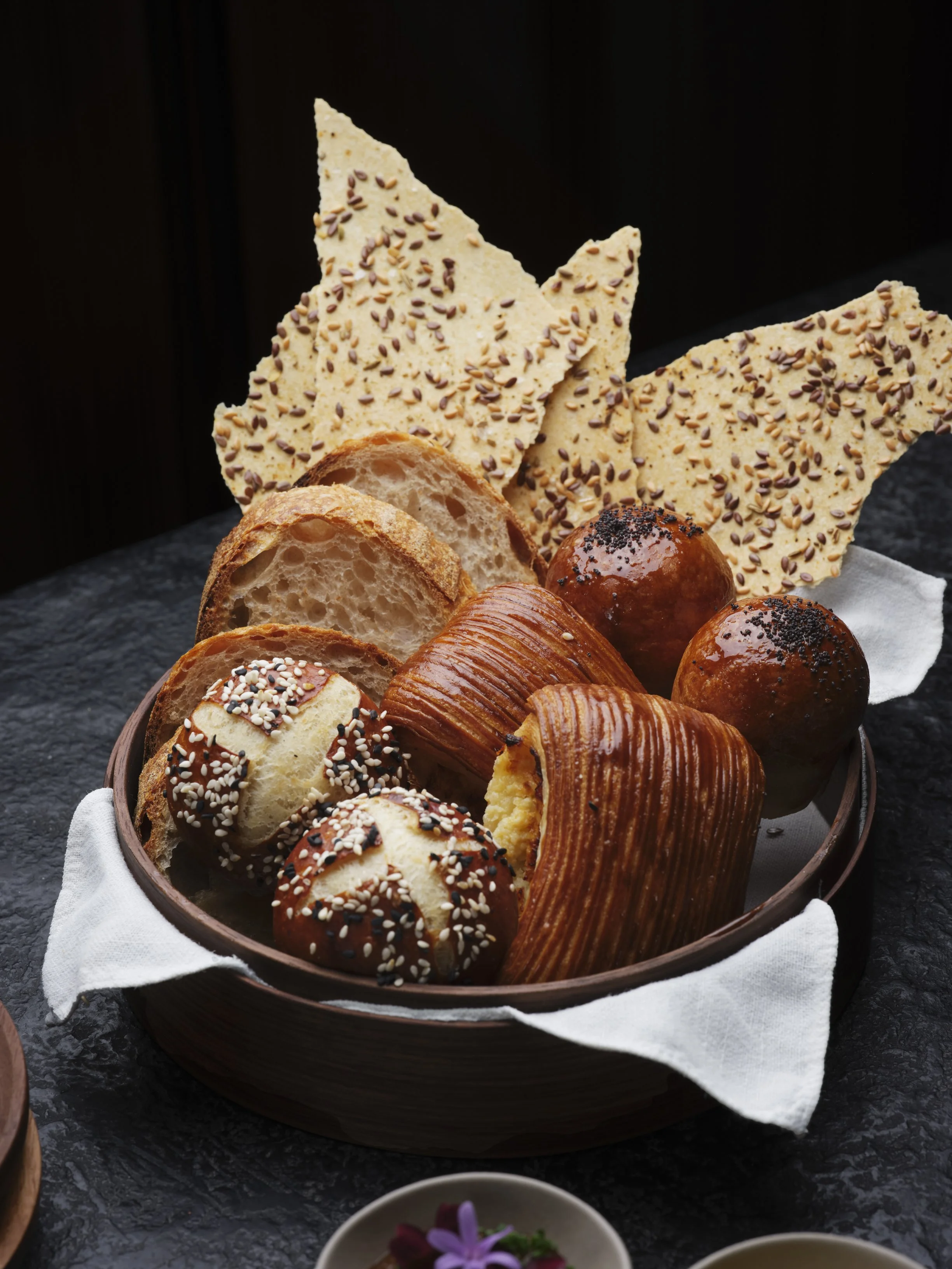 Assorted bread and crackers in a wooden bowl on a white cloth, with a small dish with purple flowers in front, on a black textured surface.