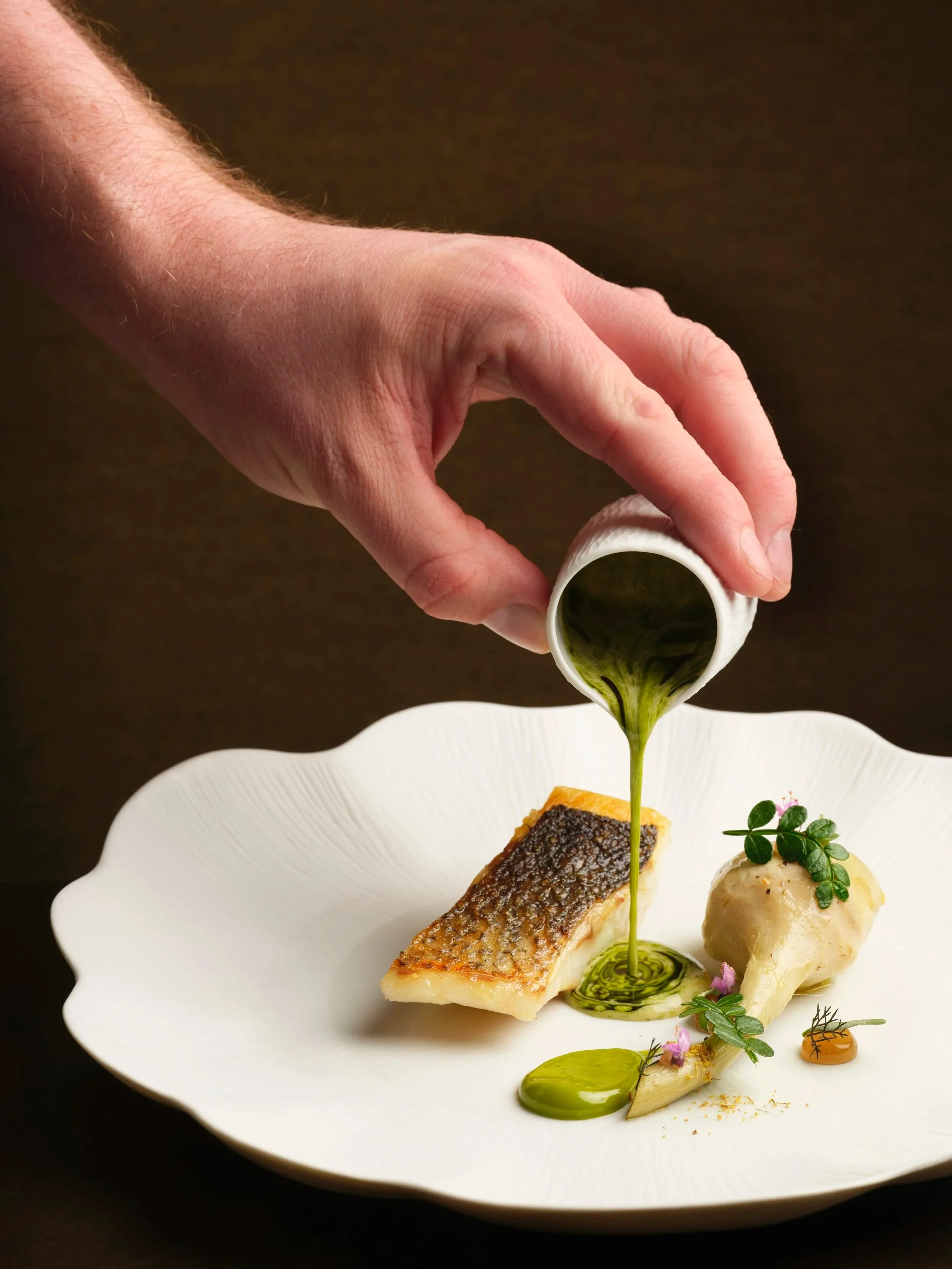 Close-up of a hand pouring green sauce over a plated gourmet dish, which includes a piece of cooked fish with crispy skin, a round scoop of mashed potatoes, and various garnishes and sauce dots on a white scalloped plate.