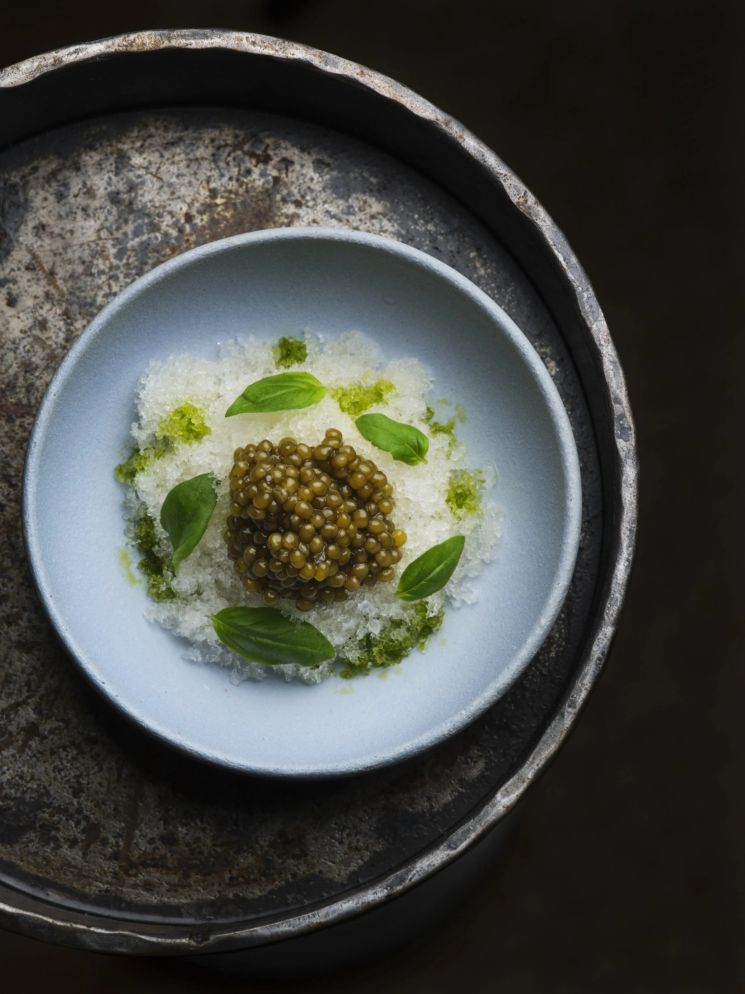 A bowl of shaved ice topped with caviar, fresh basil leaves, and green syrup, placed on a rustic metal tray.