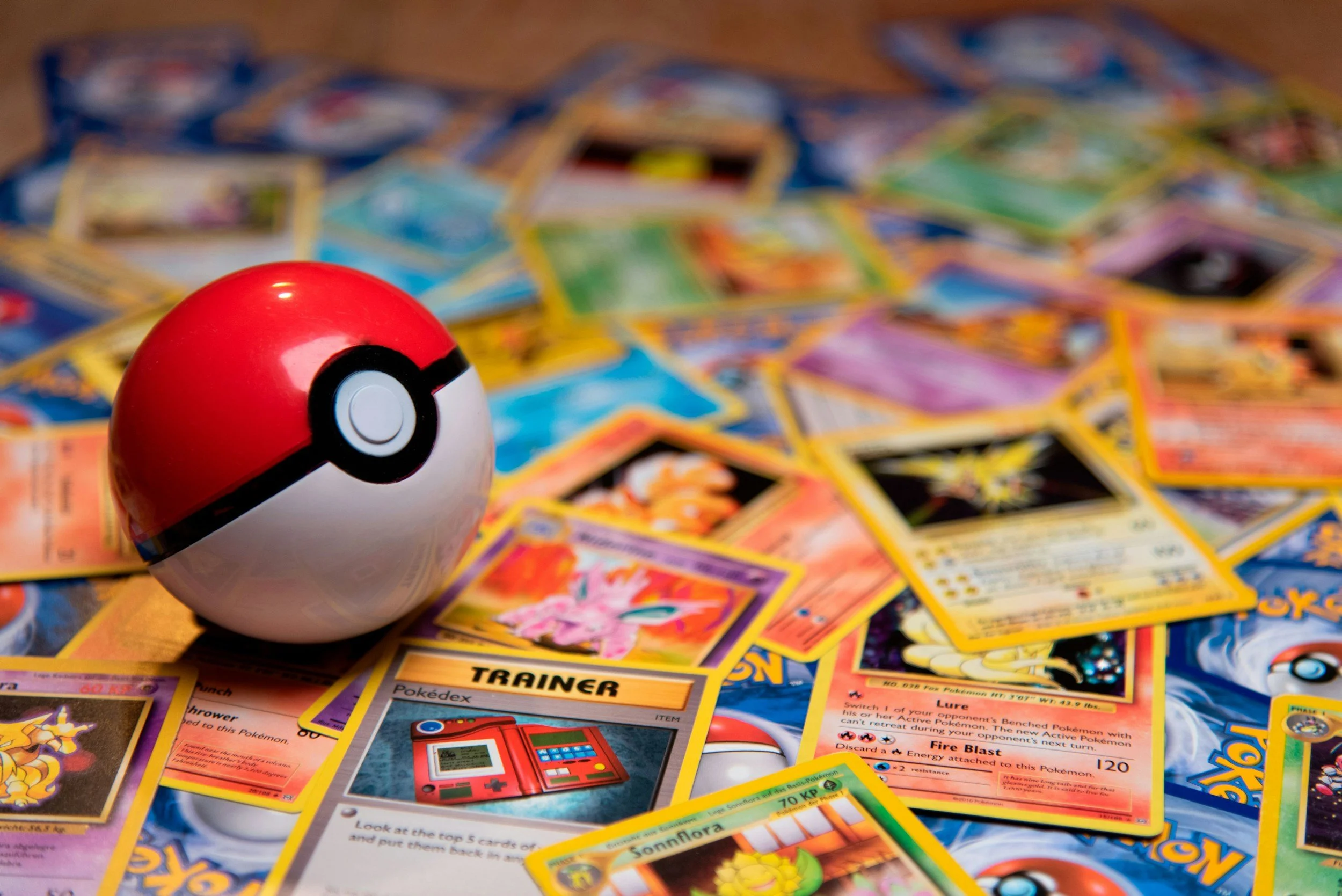 A close-up of a Poké Ball in front of scattered Pokémon trading cards on a table.