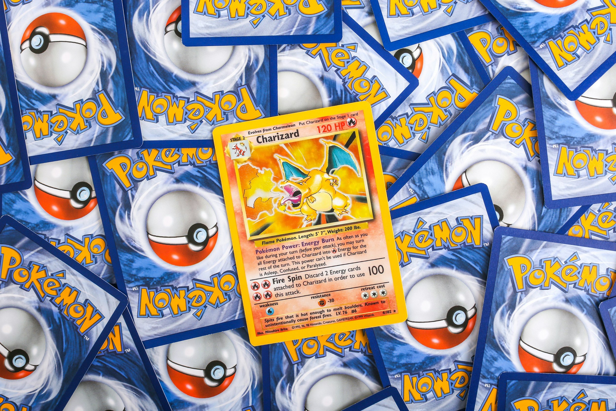 A pile of overlapping Pokémon trading cards with a focus on a Charizard card featuring an illustration of a yellow, orange, and black dragon breathing fire. The cards have the classic blue and white back design with a Poké Ball in the center.