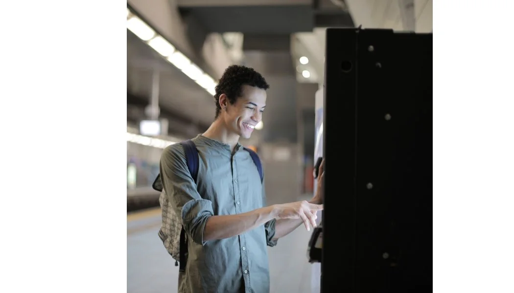A young man with curly hair and a backpack using a touchscreen at a train or subway station.