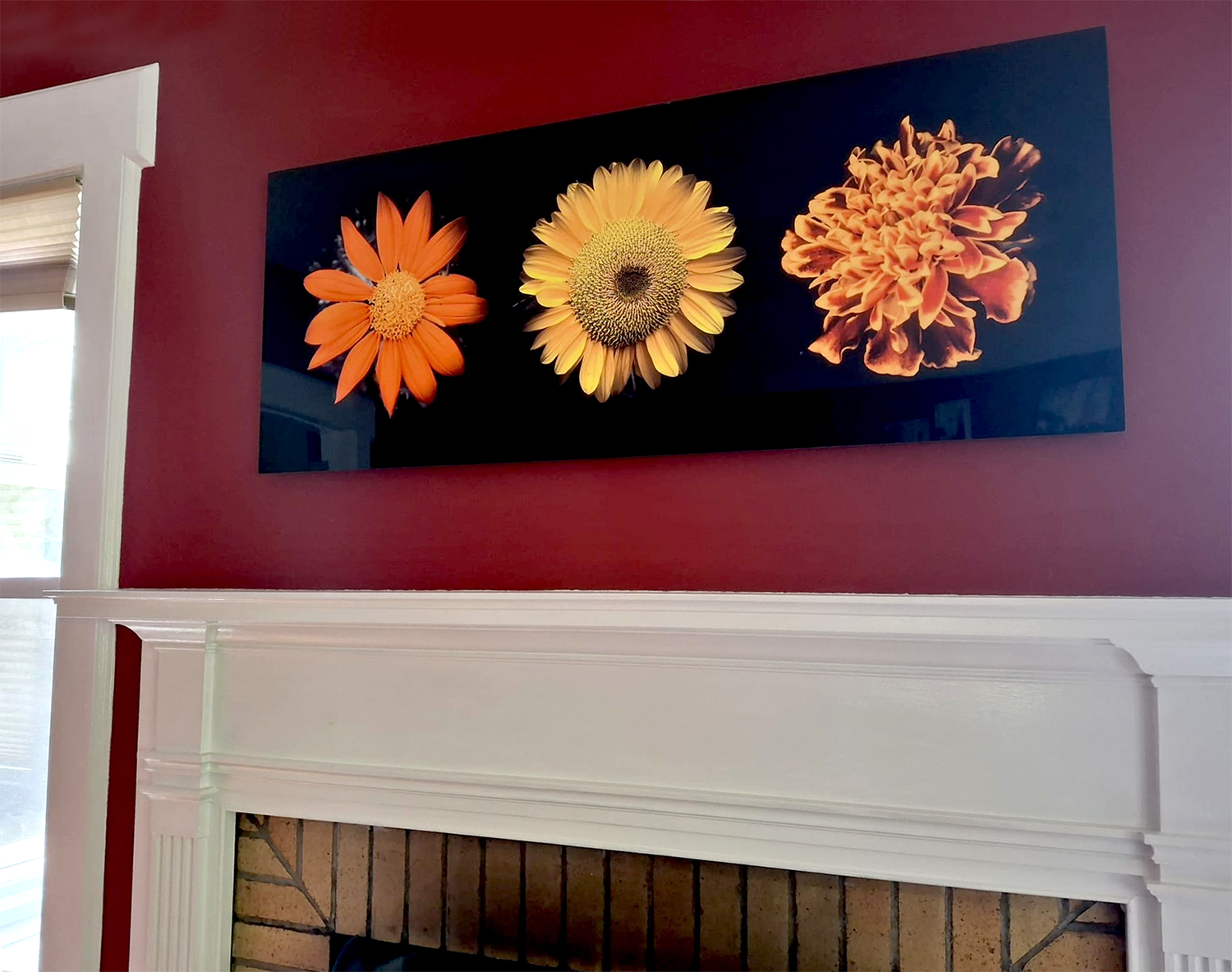 Wall art featuring three large, vibrant, orange and yellow flowers hung above a fireplace with white trim, red wall, and a window with blinds on the left side.