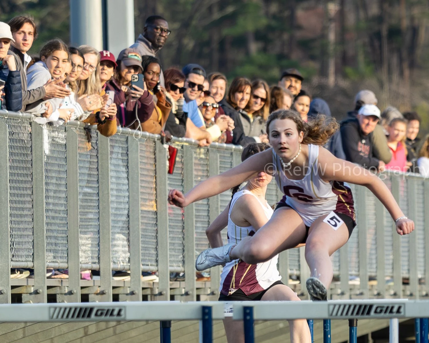 A female athlete jumping over a hurdle at a track and field meet, with spectators watching behind a metal fence.