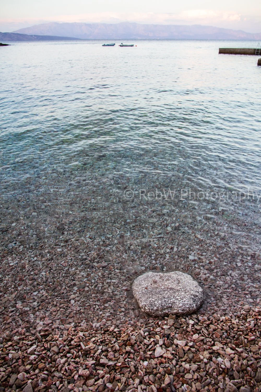 Calm lake with small rocks along the shoreline, two boats floating on the water, distant mountains in the background, and cloudy sky.