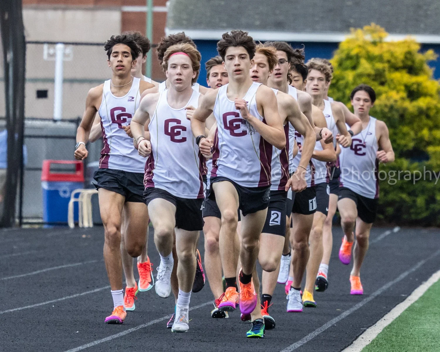 A group of young male athletes running on a track during a race, wearing white and maroon athletic uniforms with the letters 'GC' on the front.