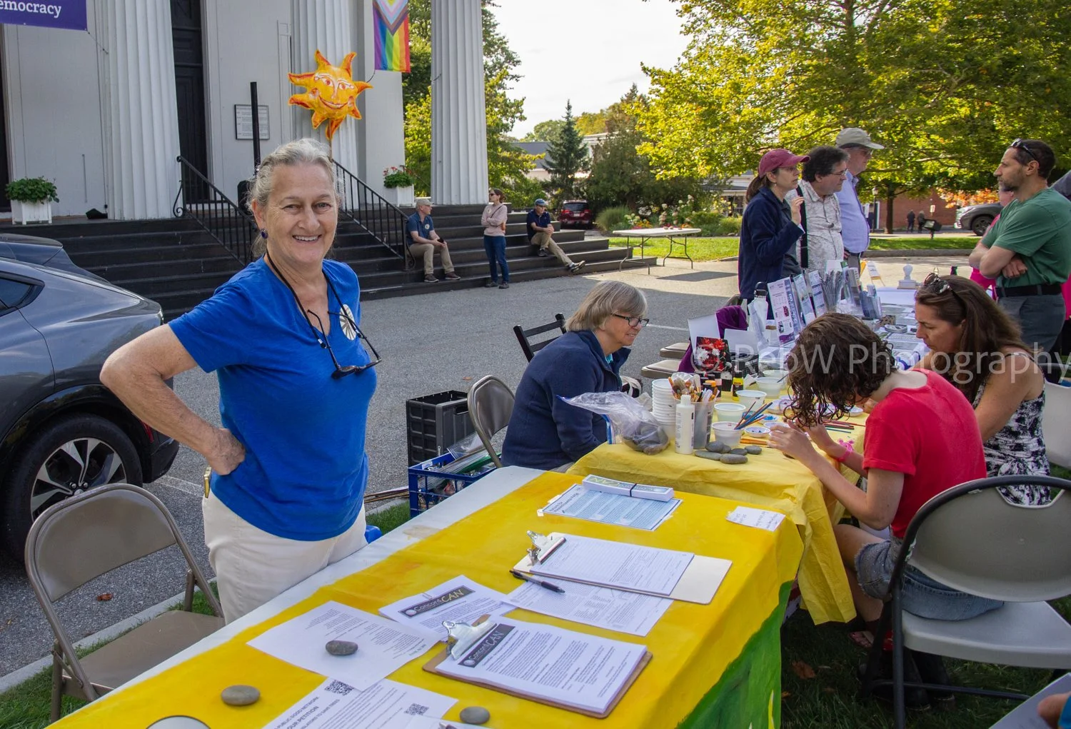 People gathered at an outdoor event in front of a building with columns, with a woman in a blue shirt smiling at the camera near a table covered with informational materials, and others engaging at a booth with papers and art supplies.