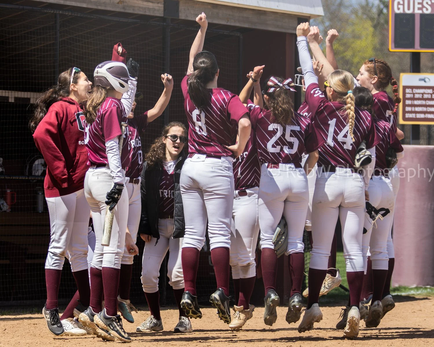 A women's softball team celebrating on the field with players gathered in a huddle, some raising fists, during a game.