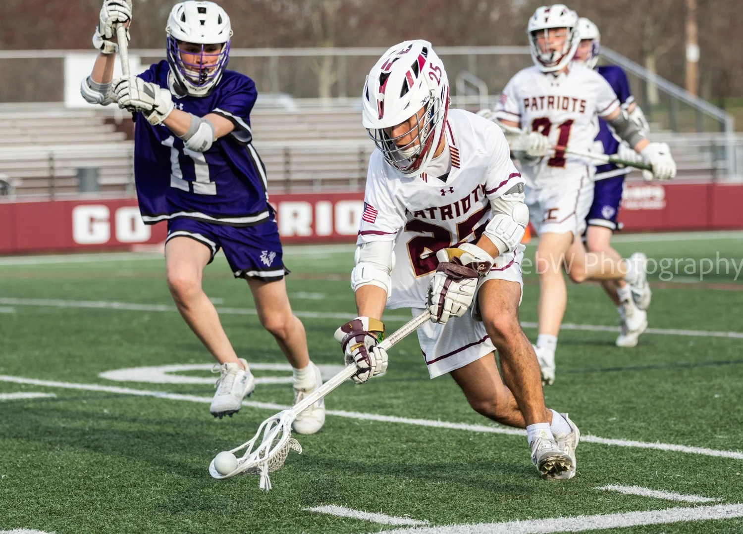 Lacrosse players in action during a game on the field, with one player in a white jersey bending down to pick up the ball with his stick, while players in purple jerseys are nearby.