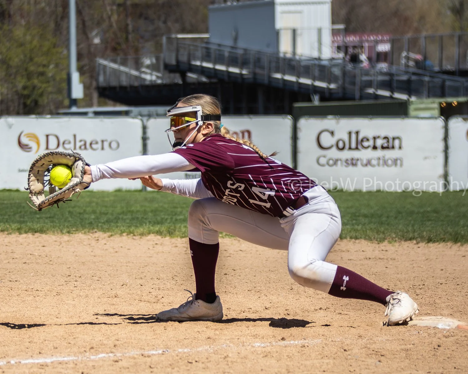 A female softball player in a maroon and white uniform crouches on a dirt field, reaching out with her glove to catch a yellow softball.