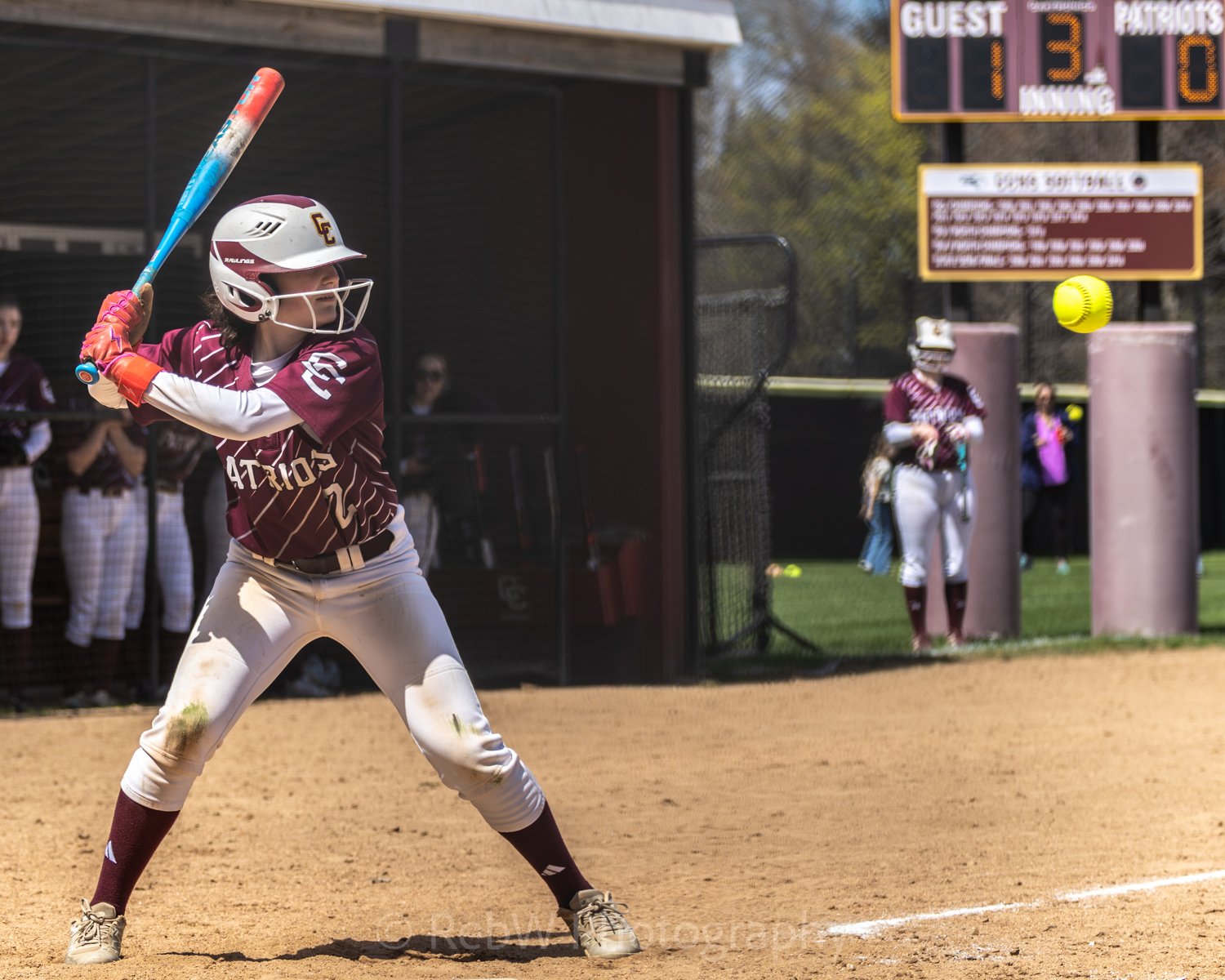 Softball player in maroon uniform prepares to swing an incoming yellow softball. Teammates are seen standing in the pin behind her.