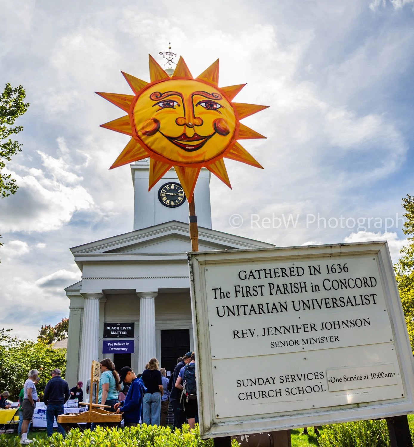 A large bright cheery sun (artwork) hovers over a sign identifying the church in the background as “The First Parish of Concord Unitarian Universalist.” A crowd of people can be seen gathered by the church.