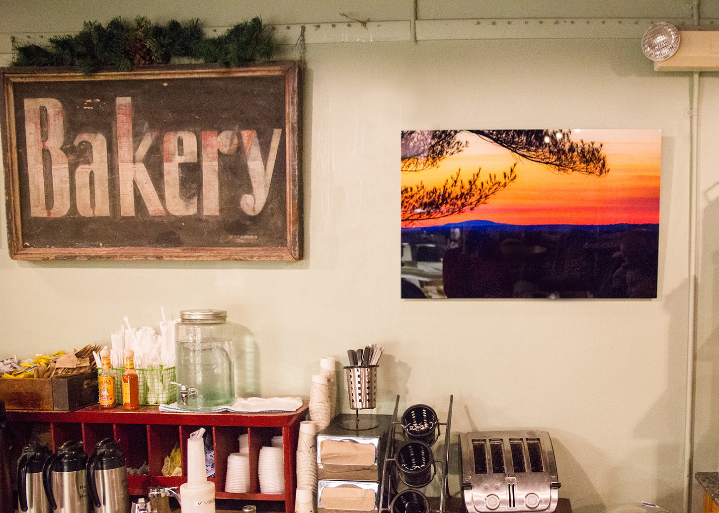 Coffee station with a chalkboard sign reading 'Bakery,' a sunset photo on the wall, and various coffee supplies including cups, straws, bottles of syrup, and toasters.