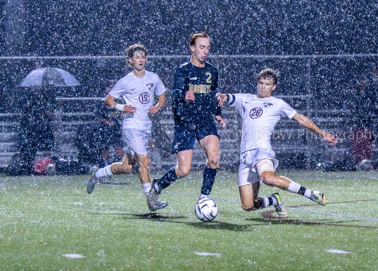 Soccer players competing in a rainy match, with one player in a dark uniform dribbling the ball while two players in white uniforms attempt to block him. One player in white is in the middle of sliding to tackle.