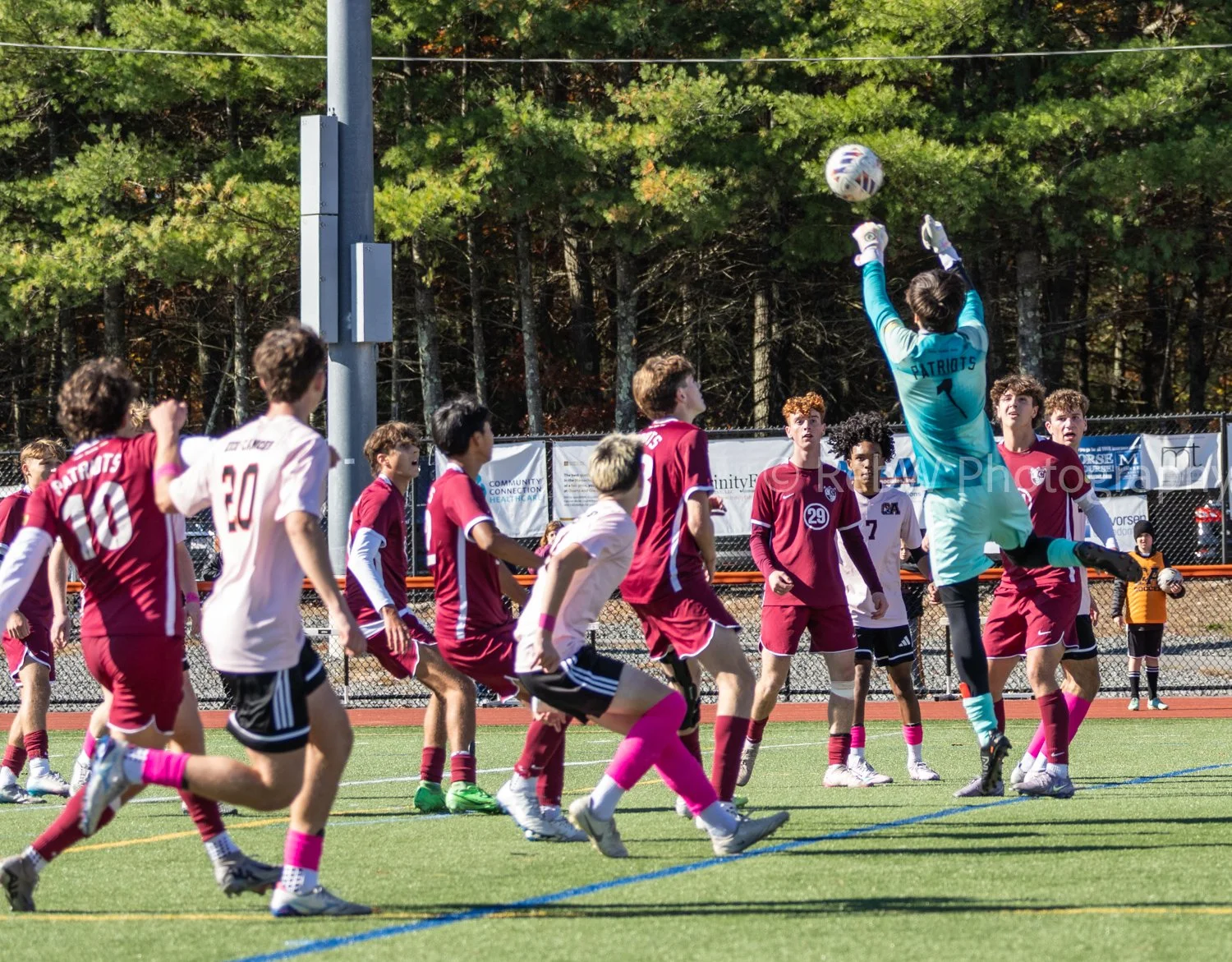 A youth soccer match showing a goalkeeper in blue jersey diving to catch the ball while other players in maroon and white jerseys watch on. The game is outdoors on a green field with a forest background.