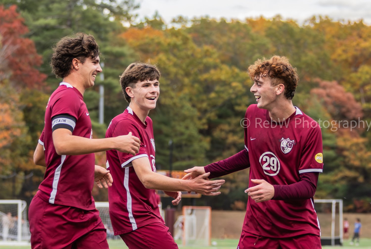 Three young male soccer players in maroon Jerseys celebrating on the field during daytime, with one wearing the captain armband, smiling and shaking hands, trees with autumn foliage in the background.