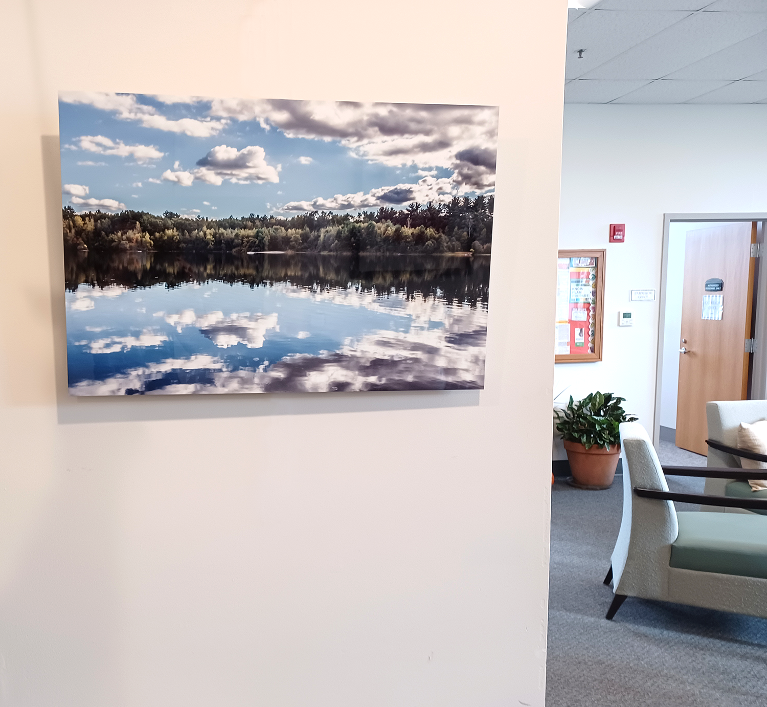 Photograph of a large landscape painting of a lake with trees and a partly cloudy sky, hanging on a white wall in a waiting room or office space.