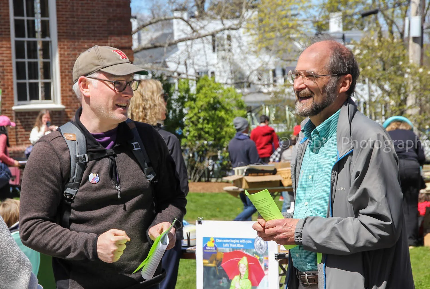 Two men, one in brown and the other grey and green, are talking and smiling at an outdoor event with other people and tables in the background.