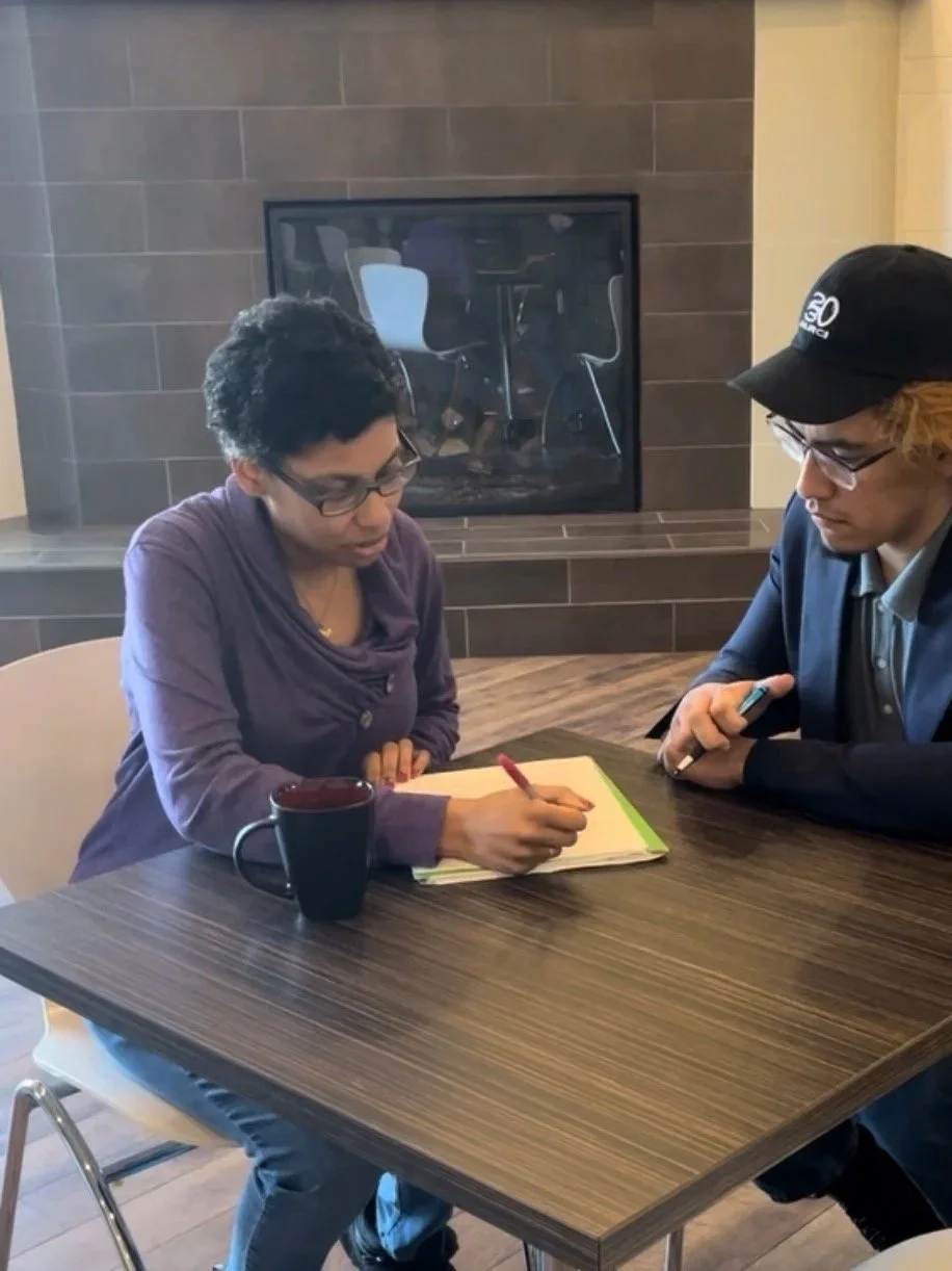 Two people seated at a table engaged in a discussion about the man's writing. The coach is providing feedback and critique.