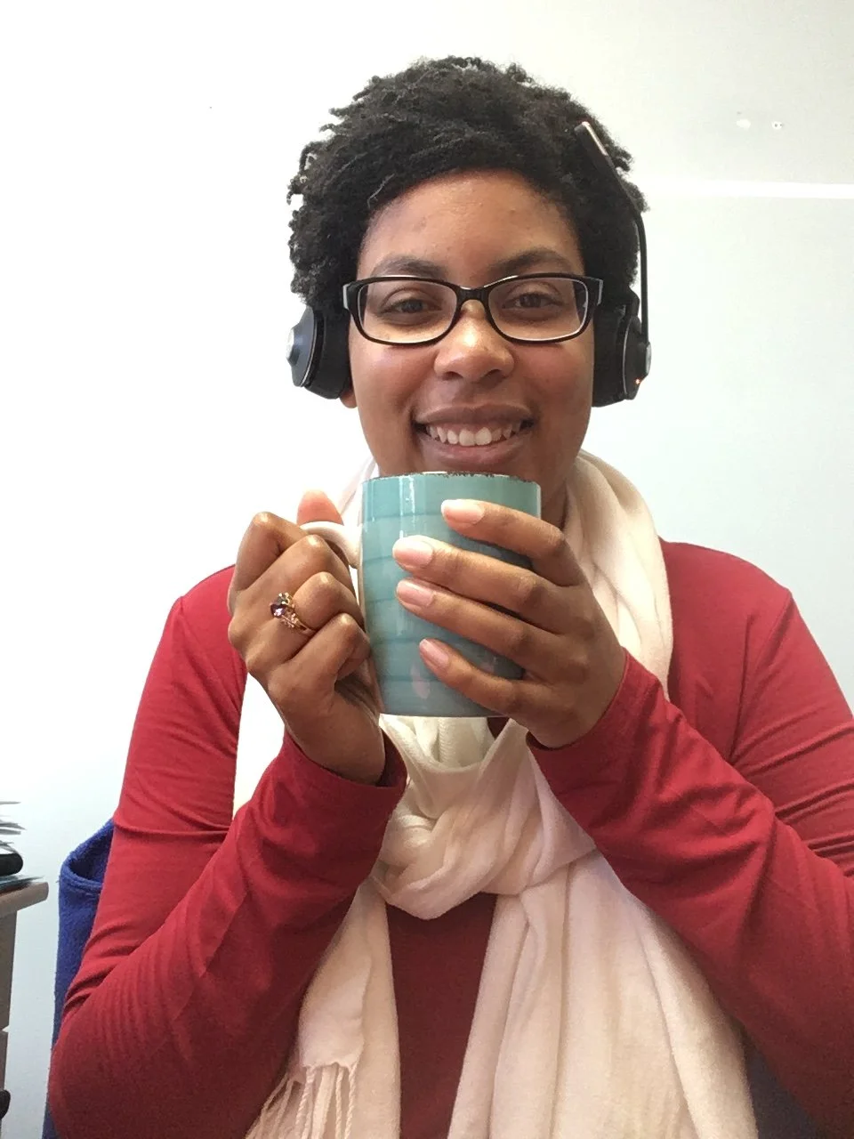Smiling woman with glasses and headphones holding a mug in an office setting. She is ready to help guide you in the writing and publishing process.