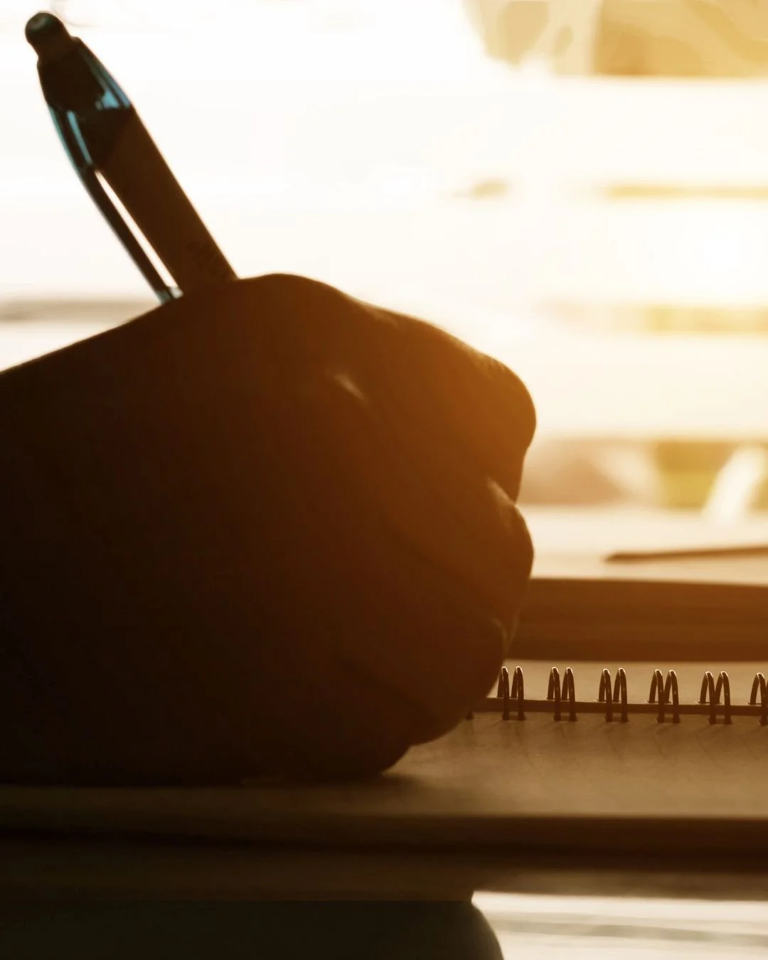 A person writing in a notebook with a pen, backlit by sunlight coming through a window.