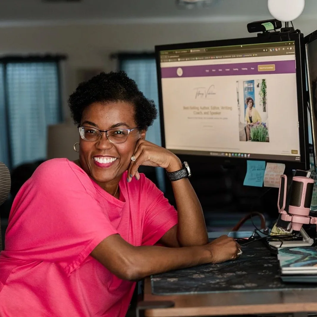 A woman with short curly hair, glasses, and a bright pink shirt smiling at the camera while sitting at a desk equipped with a computer, microphone, and notebooks in a home office. She is ready to edit her next manuscript.