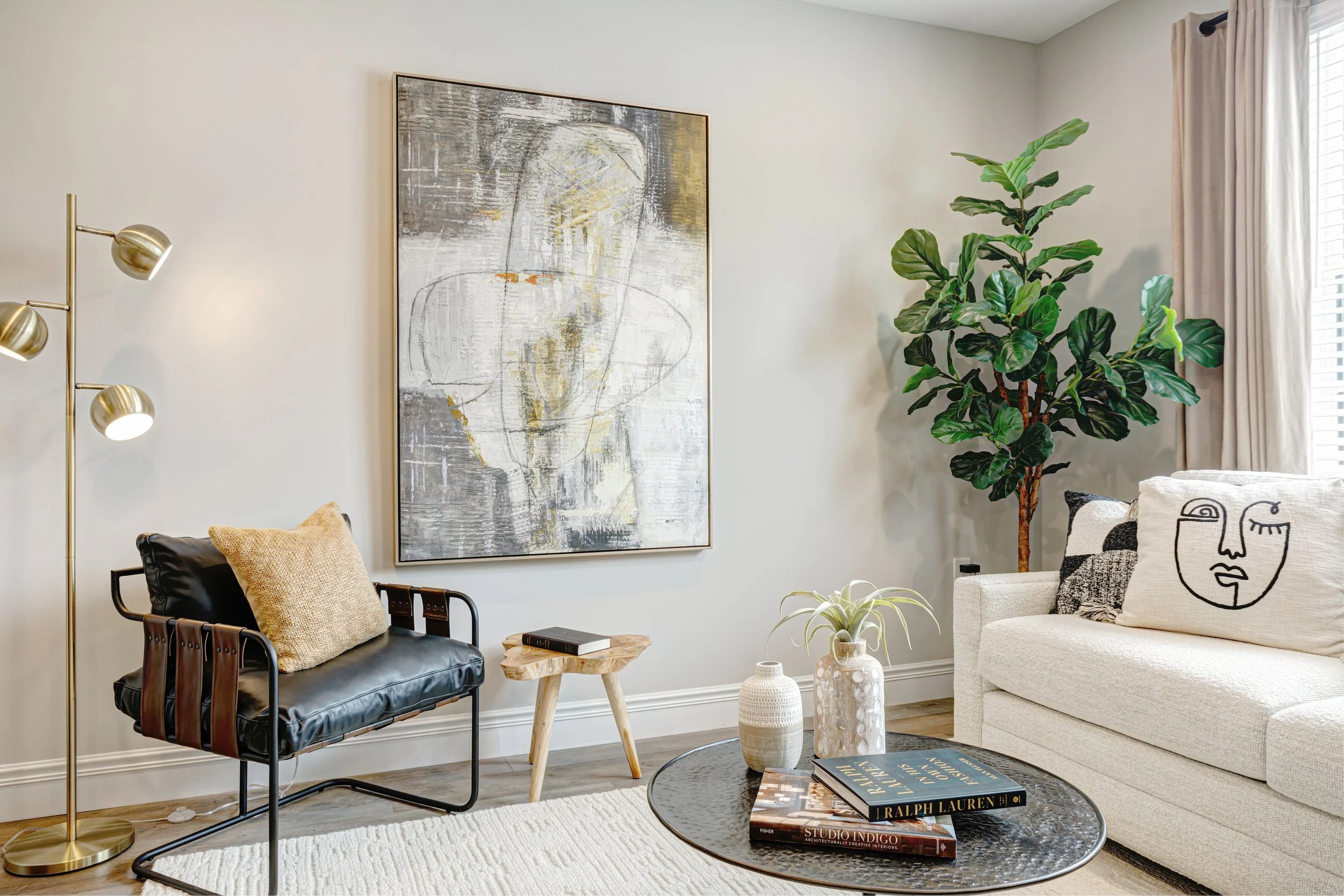 A modern living room corner with a black leather chair with a beige pillow, a small wooden side table, a tall gold floor lamp with three adjustable heads, a large abstract art print on the wall, a tall potted plant, a white upholstered sofa with blac