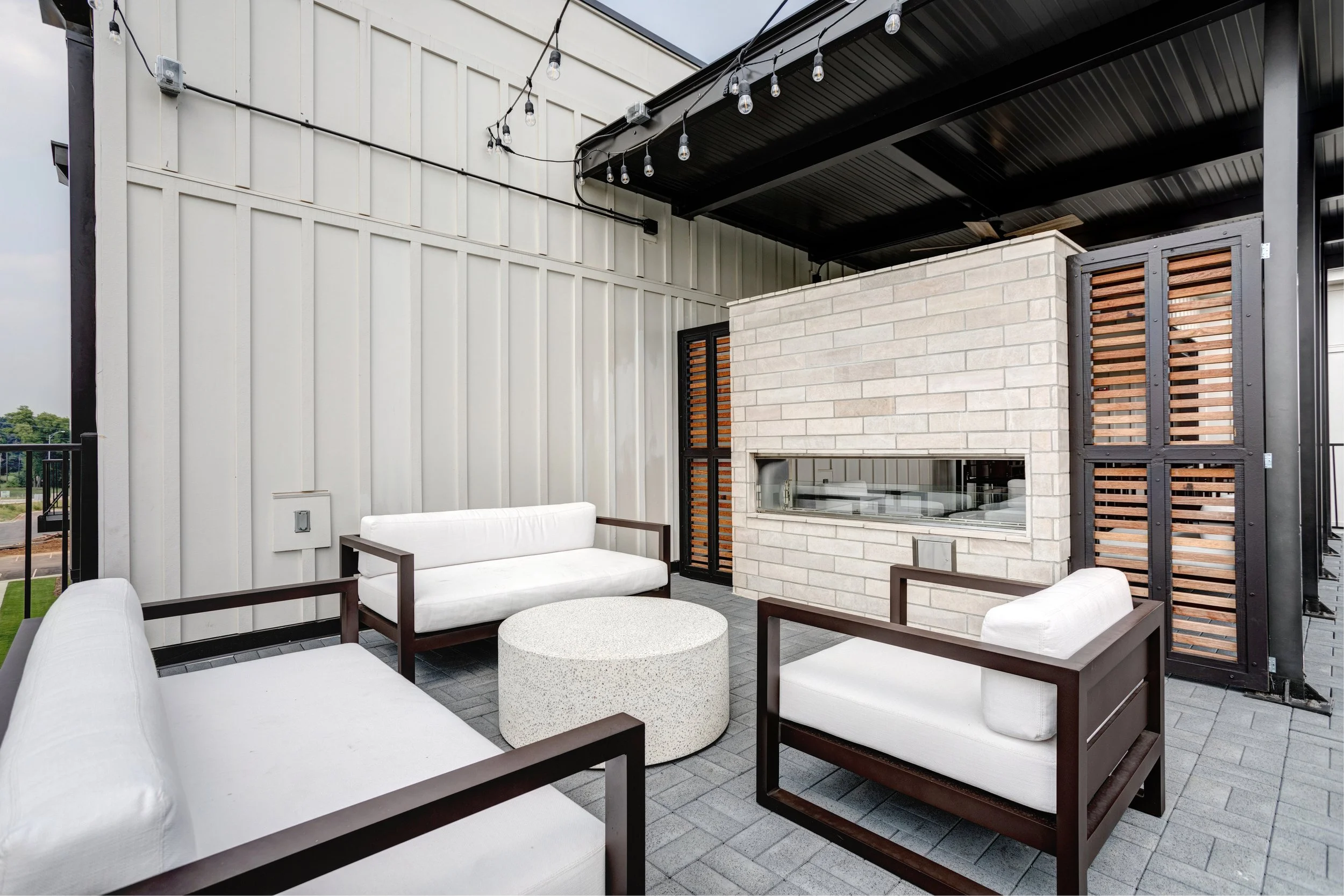 Outdoor patio with white cushioned seating, a round coffee table, a brick fireplace, and string lights hanging from the black ceiling. Modern design with a mix of metal, brick, and wood materials.
