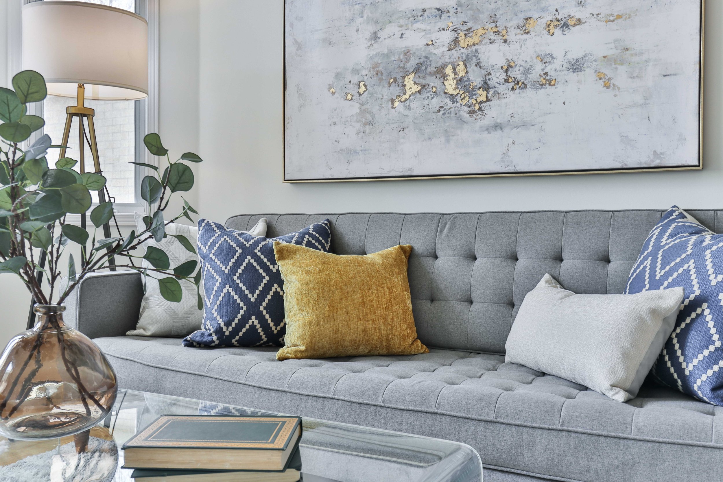 A living room with a gray tufted sofa decorated with multiple pillows in blue, yellow, and white patterns. On the left, a glass table holds a book and a decorative glass vase with green leaves. Behind the sofa, a large abstract painting with gold, gray, and white hues hangs on the wall. To the left, a tall wooden lamp with a white shade stands next to a window with white trim, letting in natural light.