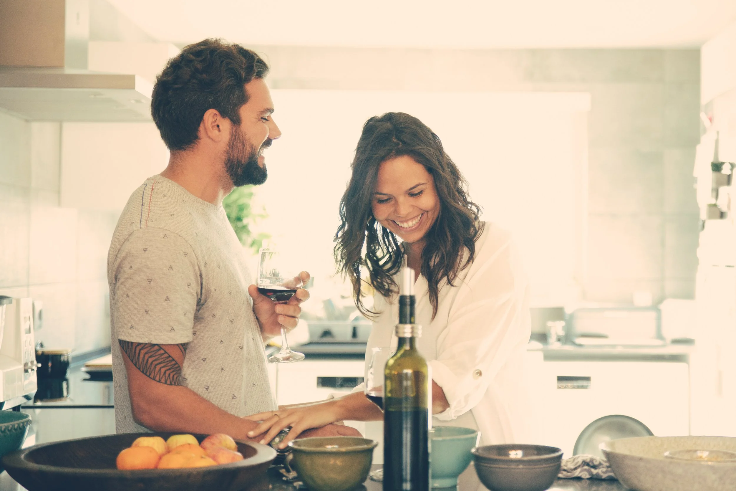 A man with dark hair, beard, and a tattoo on his arm, holding a glass of red wine, standing next to a woman with long curly hair, smiling and looking down, in a brightly lit modern kitchen.