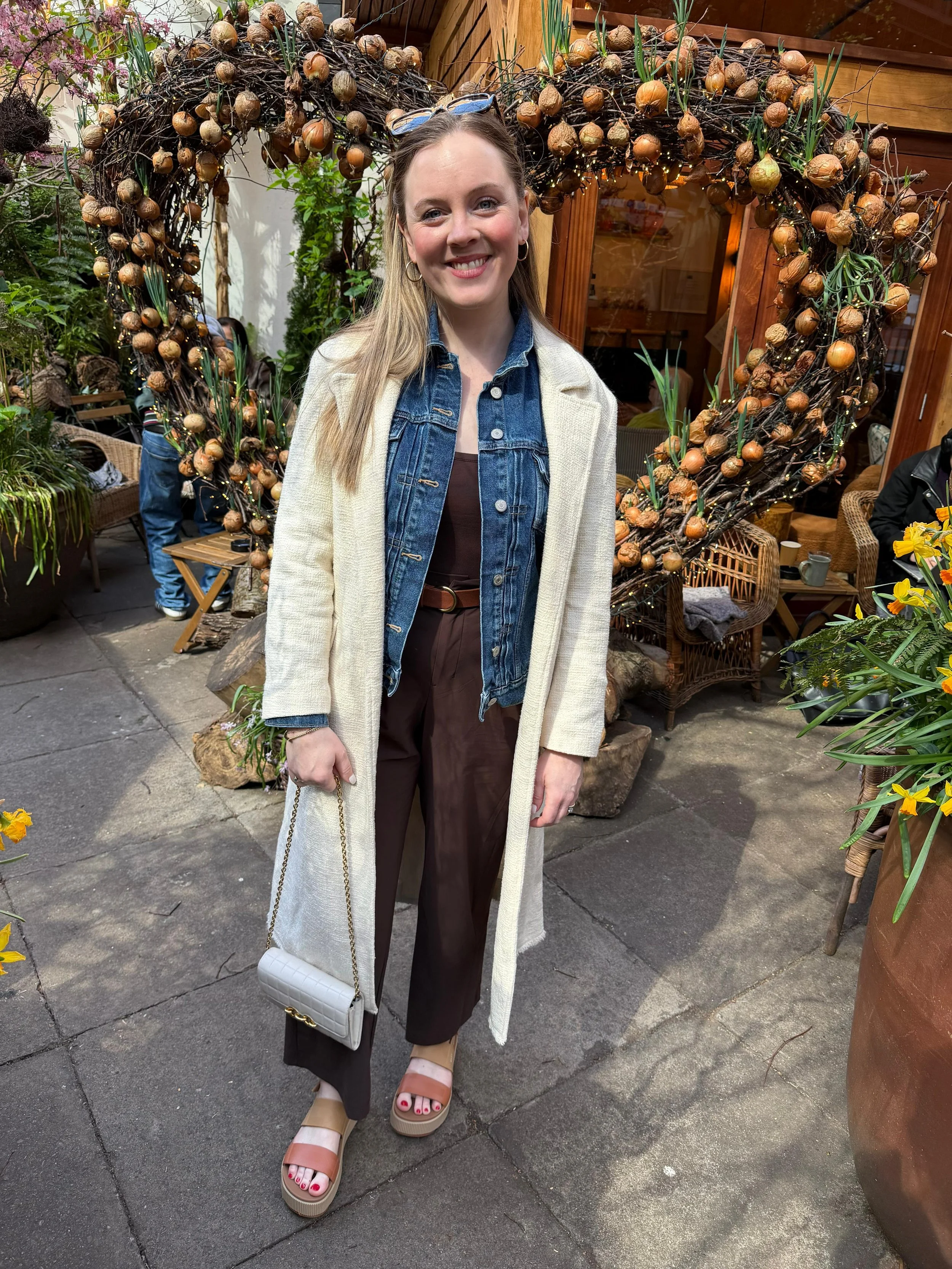 A woman with long blonde hair and earrings smiling at the camera standing in a garden or patio area with decorative plant arrangements. She is wearing a denim jacket over a brown top, dark brown pants, and beige and pink sandals. She holds a white quilted handbag with a gold chain strap.
