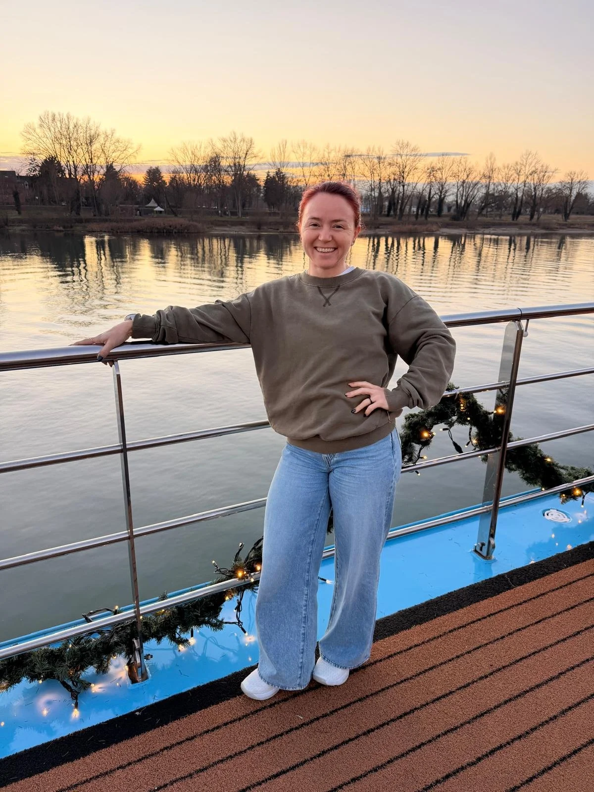 A woman standing on a boat deck near a body of water during sunset, smiling, dressed casually in a gray sweatshirt and jeans, with holiday decorations on the railing.