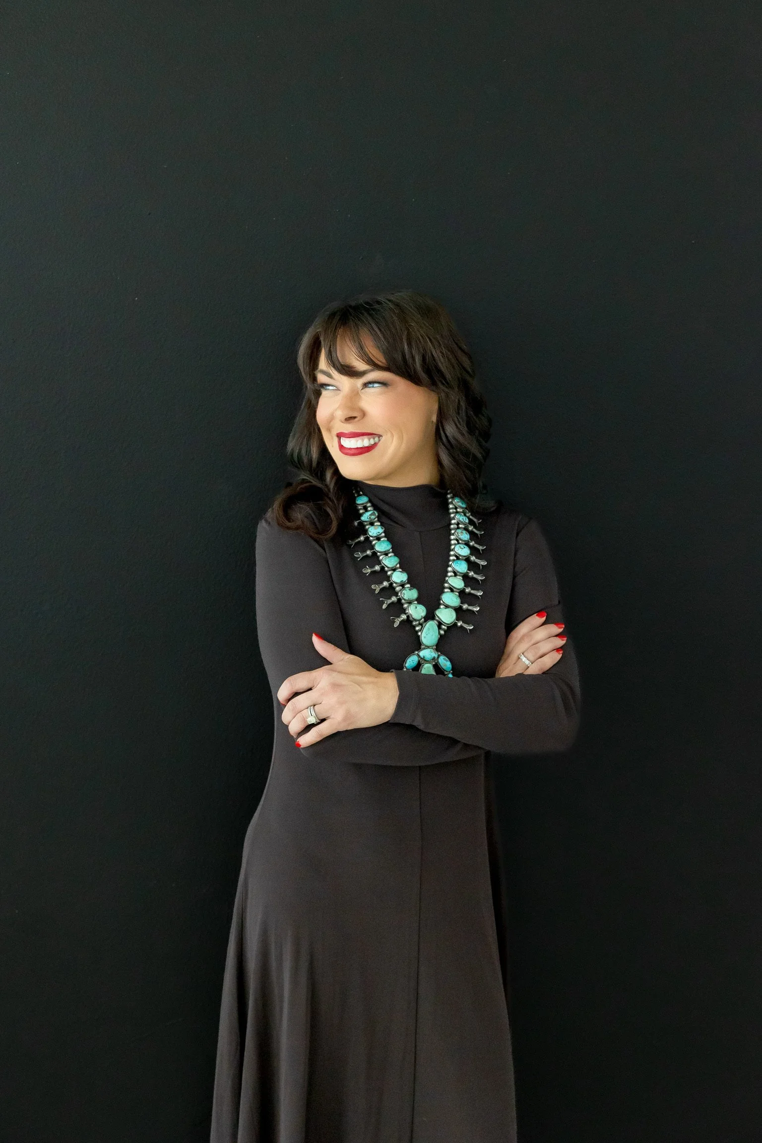 A woman with dark brown hair styled in loose curls, wearing a long black dress with long sleeves, red lipstick, and red nail polish, standing against a plain black background. She is smiling and looking to her right with her arms crossed, accessorized with turquoise jewelry, including a necklace and ring.
