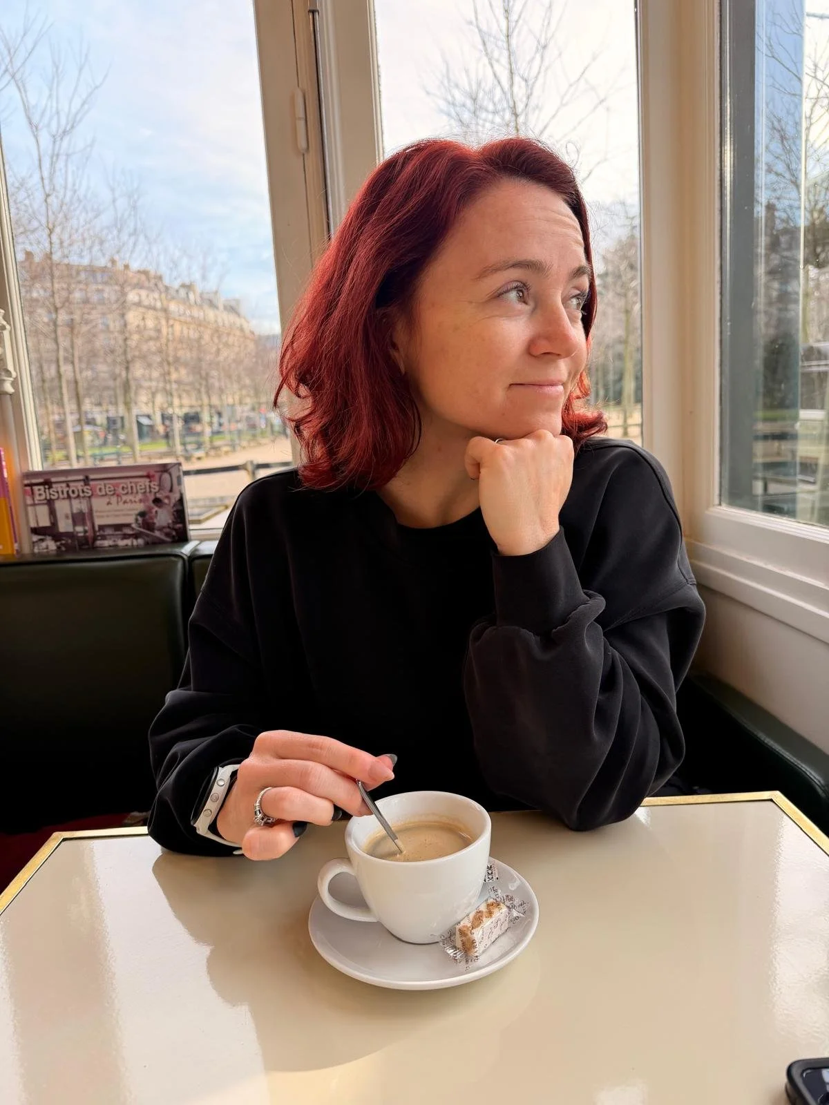 A woman with red hair sits at a table in a cafe, holding a spoon and a cup of coffee, gazing out the window.