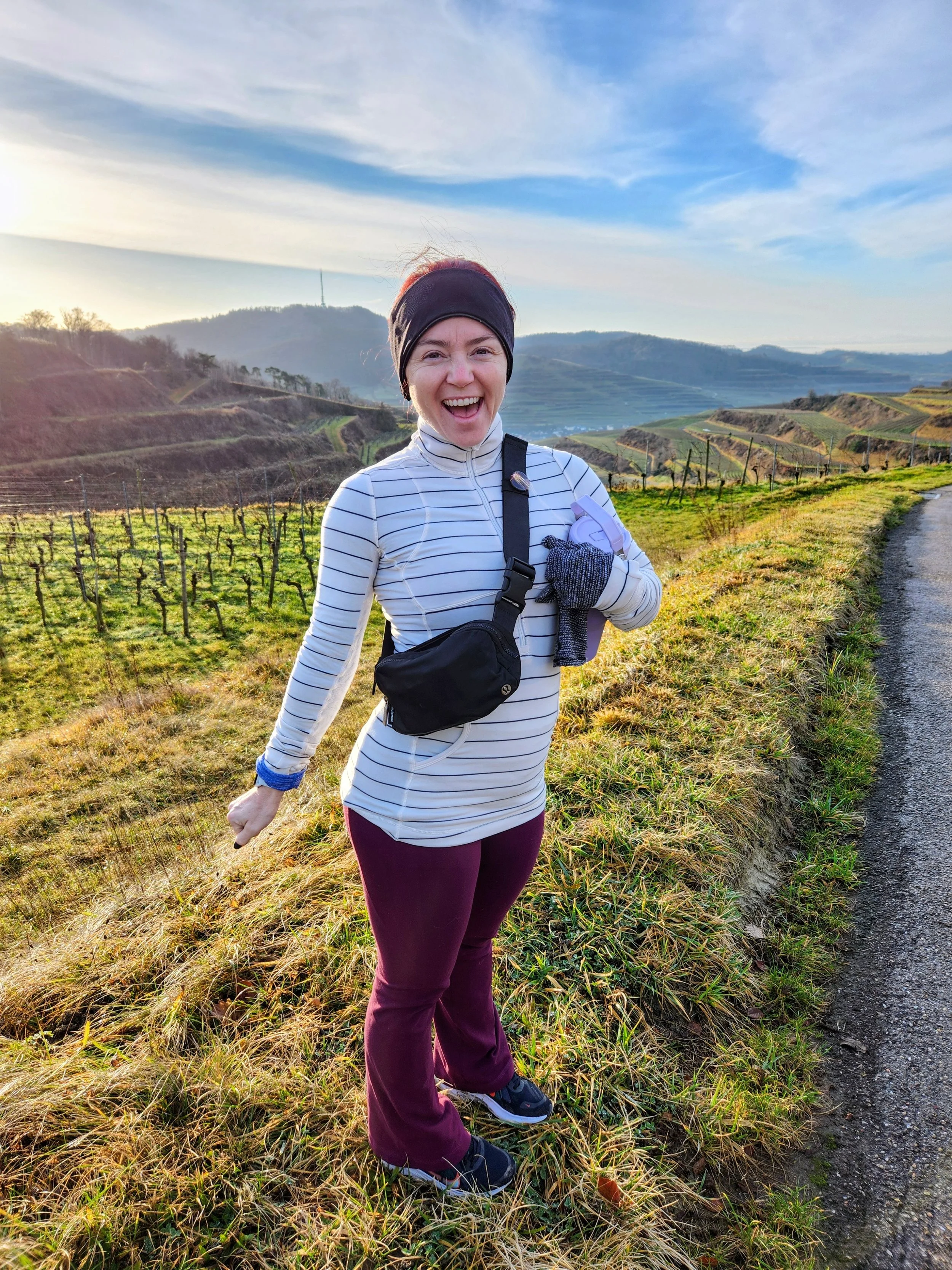 A woman in athletic clothing smiling and enjoying a hike in a rural landscape with rolling hills, vineyards, and a partly cloudy sky during sunset.