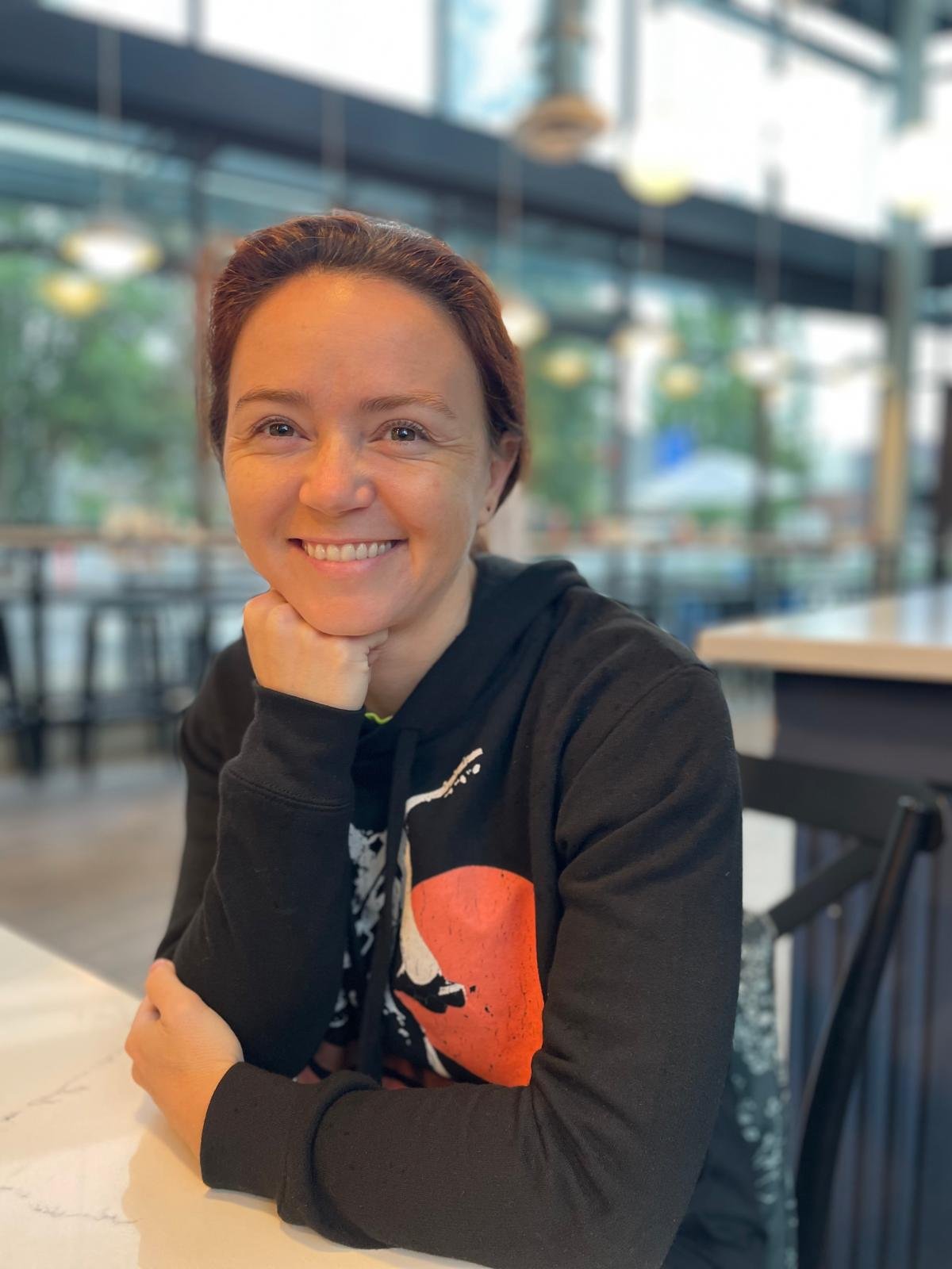 A woman with short reddish hair smiling while sitting at a table in a modern, well-lit café or restaurant with large windows and blurred background of trees and outdoor setting.