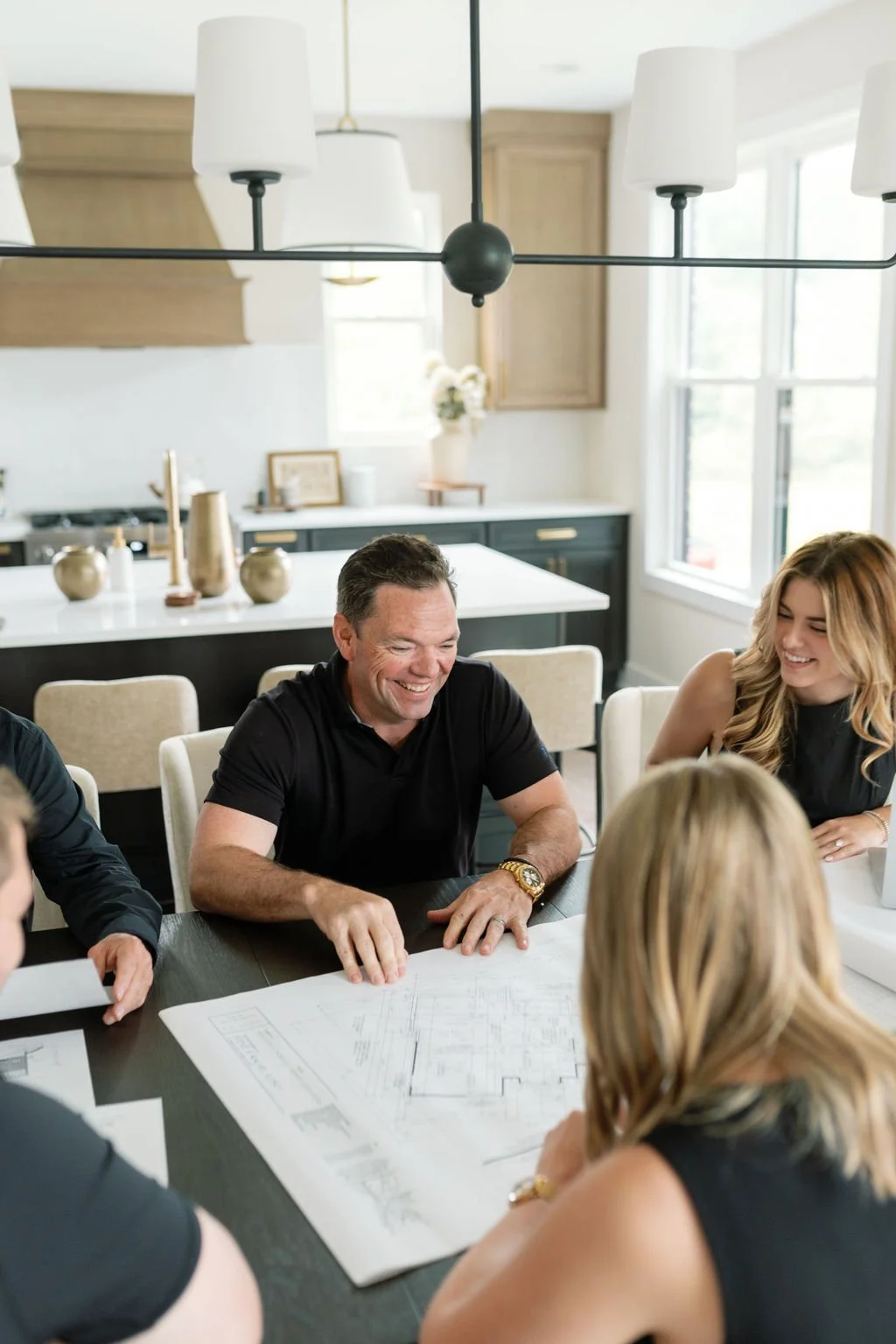 Group of people smiling and discussing blueprints at a dining table in a  kitchen.