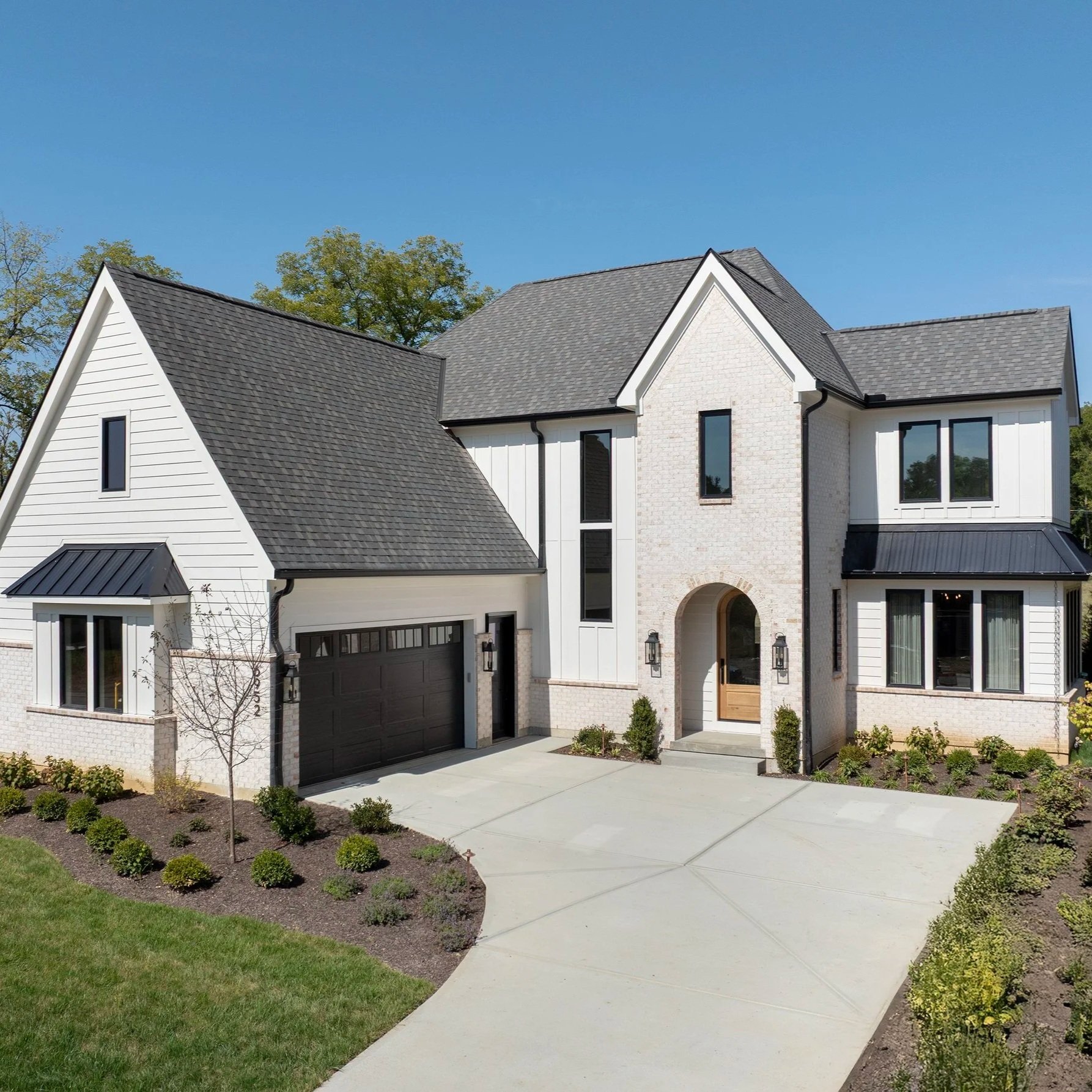 Custom two-story house with a gray roof, white siding, and large windows, surrounded by a landscaped yard with a young tree and green shrubs, and a concrete driveway leading to a garage and front door, under a clear blue sky.