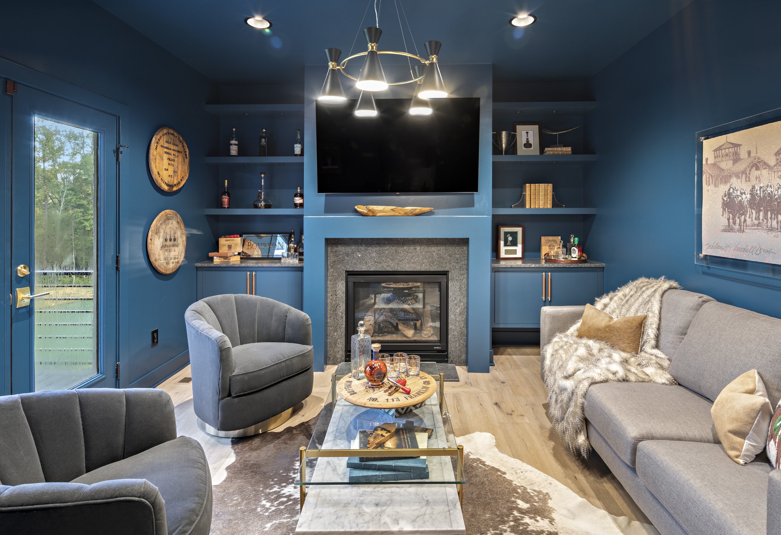 A cozy living room with dark blue walls, a fireplace, and built-in shelves. There are gray and dark-colored sofas, a glass coffee table with books and decorative items, and a large TV mounted above the fireplace. The room is decorated with framed artwork, bottles of liquor on the shelves, and a patterned rug.